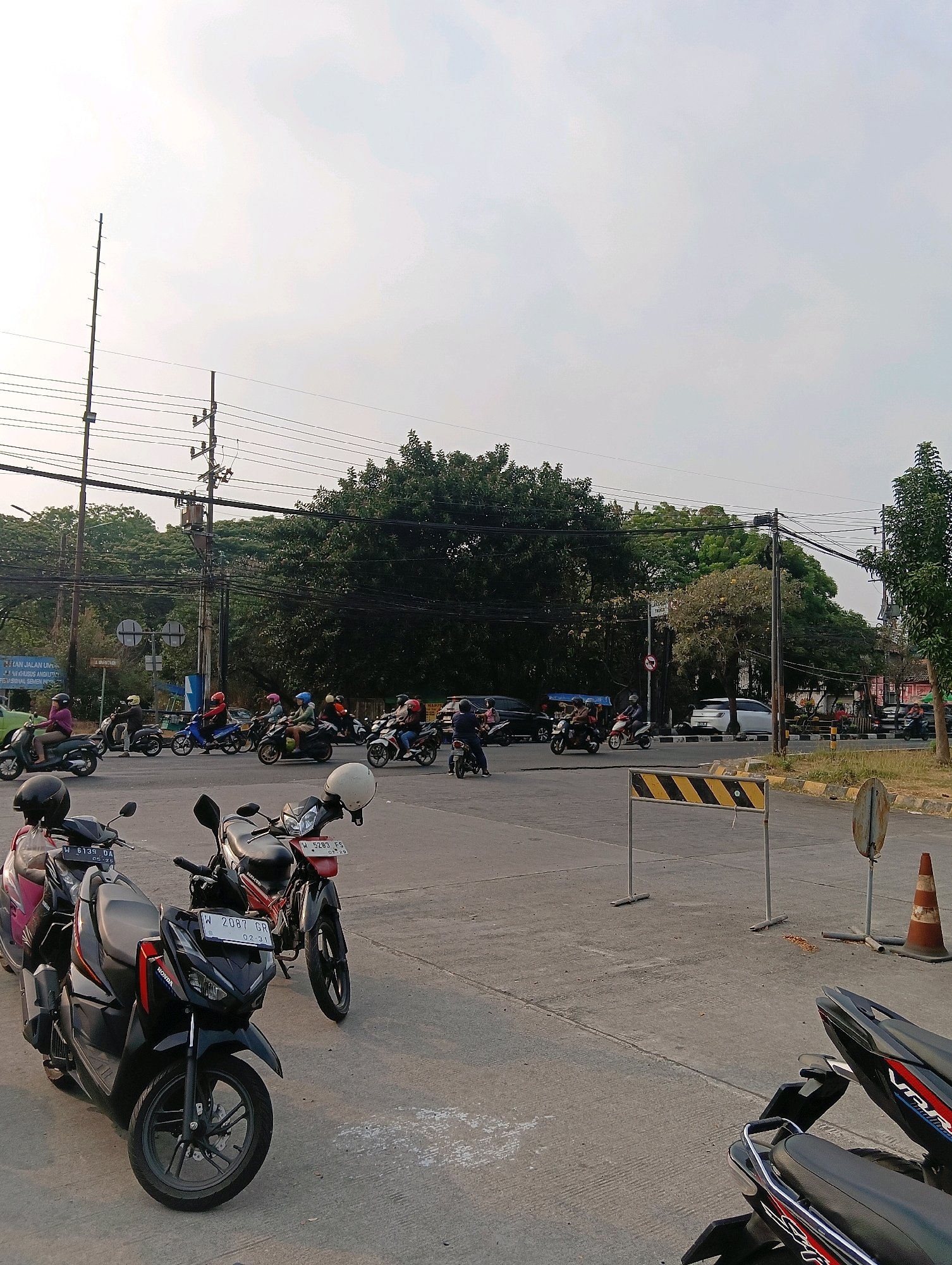 The image depicts a street scene in the city of Gapurosukolilo, Indonesia, during the day, likely late afternoon given the low angle of the sun casting long shadows. The foreground is occupied by several parked motorcycles, some with helmets placed on them. A black Honda motorcycle with red accents is prominently displayed, showing its license plate which reads "W 2087 GR". To its left, a pink motorcycle is partially visible. In the middle ground, a busy intersection is visible with numerous motorcycles and a few cars navigating the road. The riders are predominantly wearing helmets, and some are carrying passengers. The environment is characterized by lush green trees lining the street, overhead power lines supported by poles, and various traffic control signs and barriers, including a portable yellow and black striped barrier and a traffic cone. The sky is overcast with a pale, hazy quality. The overall impression is of a typical urban traffic environment in Indonesia.