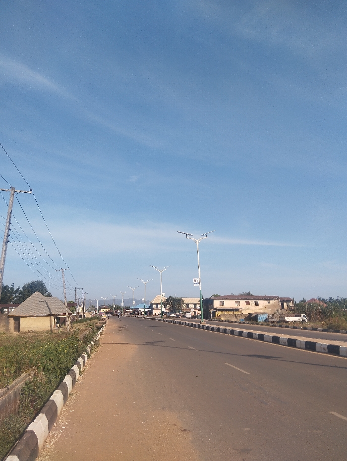 The image displays a wide, paved road in Unguwan Rimi, Nigeria, under a clear blue sky with minimal cloud cover. The road, likely a thoroughfare, features a central median delineated by black and white striped curbs. A series of modern streetlights, each with multiple curved arms, are installed along this median. On the left side of the road, a dirt shoulder with green vegetation in a ditch precedes a small, circular, mud-colored hut with a conical roof. A wooden utility pole with numerous power lines stands adjacent to the hut. Further down the left side, additional indistinct structures and a second utility pole are visible. On the right side of the road, beyond the median, a line of various buildings, ranging from multi-story structures to smaller commercial establishments, stretches into the background. A white pickup truck is discernible on the right side of the road in the distance. A lone figure, possibly on a motorcycle or bicycle, is observed in the far distance on the road. The scene is illuminated by direct sunlight, casting subtle shadows, and appears to represent a typical daytime urban environment.