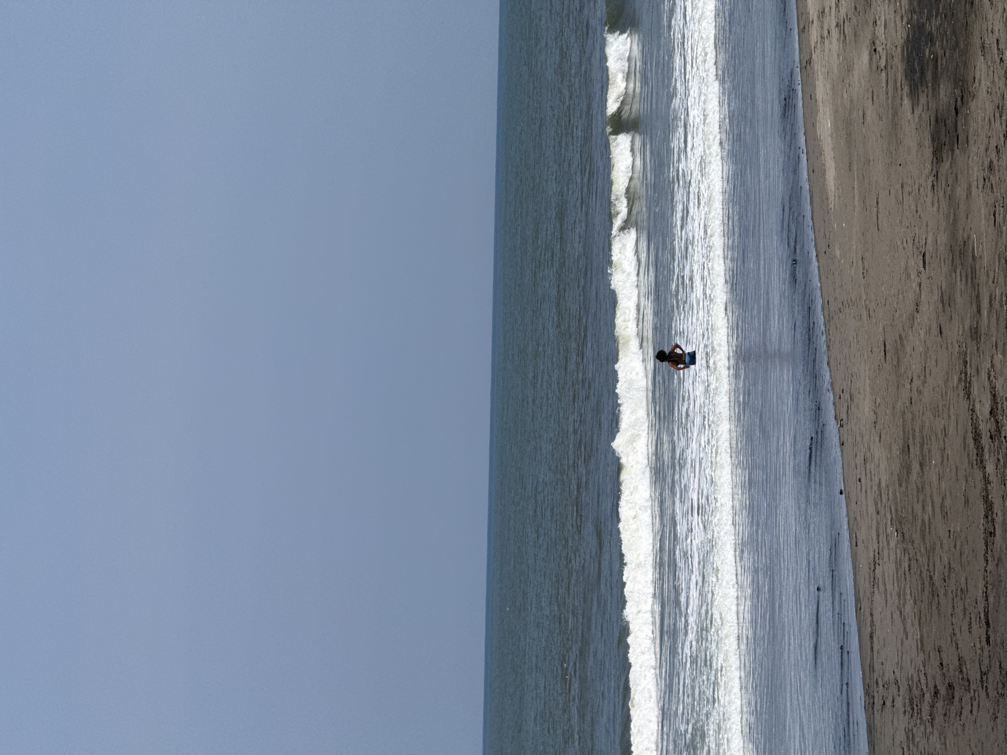 The image displays a single individual standing in the surf of a beach in Belalang, Indonesia. The person, viewed from the back, is shirtless and wears blue shorts, facing the open ocean. Breaking white waves surround the individual, whose lower legs are submerged in the water. The foreground features a dark, damp sand beach transitioning to drier sand. The midground shows the ocean with multiple wave lines, extending to a distant horizon under a clear, light blue sky.
