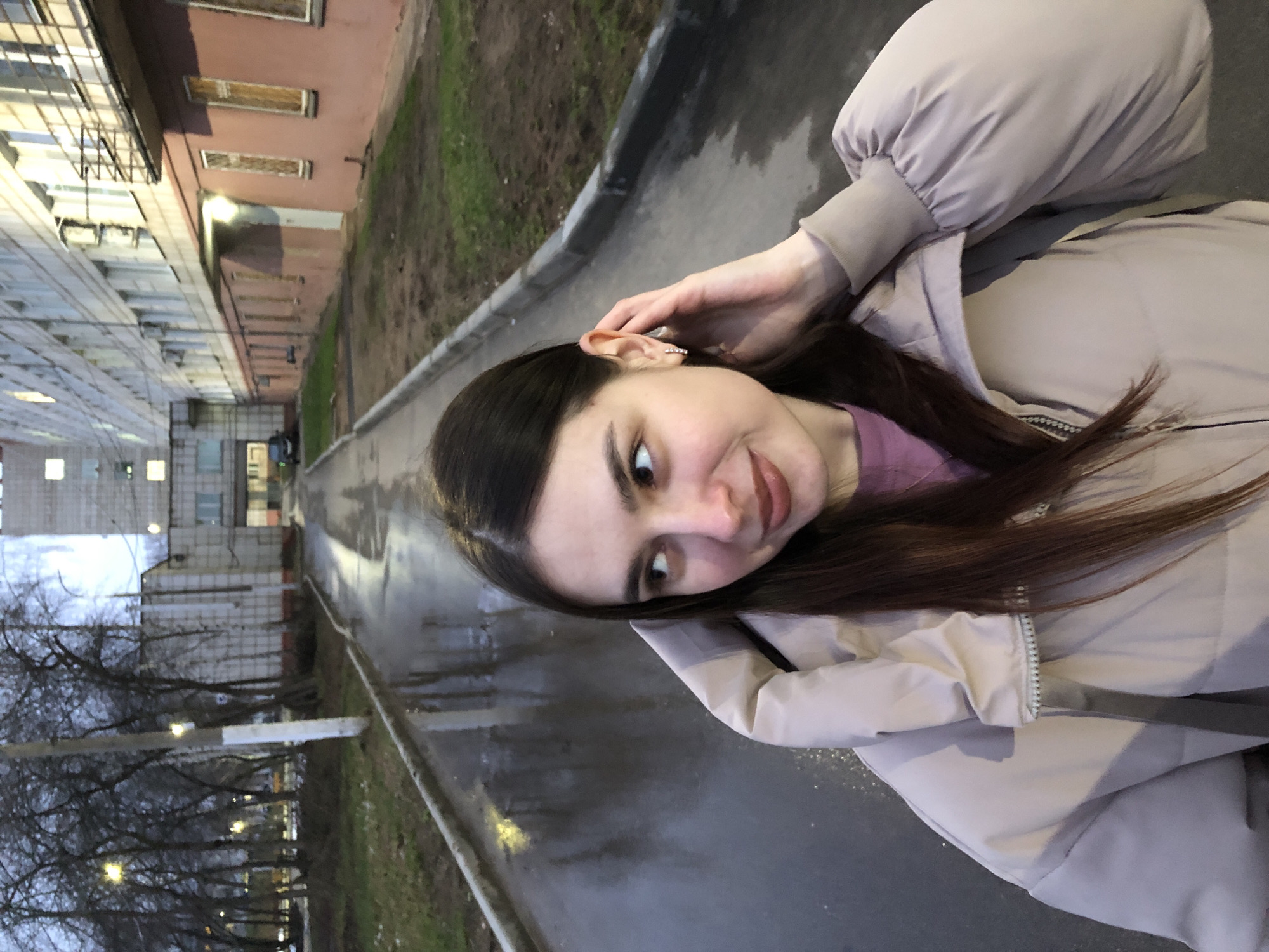 A young woman with dark hair and fair skin poses for a selfie outdoors. She is wearing a light-colored puffer jacket and a pink top. Her left hand is raised near her ear, which has a decorative earring. The background consists of a wet, grey pavement alongside a curb separating the road from a grassy area. Trees without leaves are visible along the left side of the image. Buildings, some with lit windows, are visible on both sides of the road. The buildings appear to be apartments. This photograph was taken in a city setting on what seems to be an overcast day in Chaikovsky, Russia.