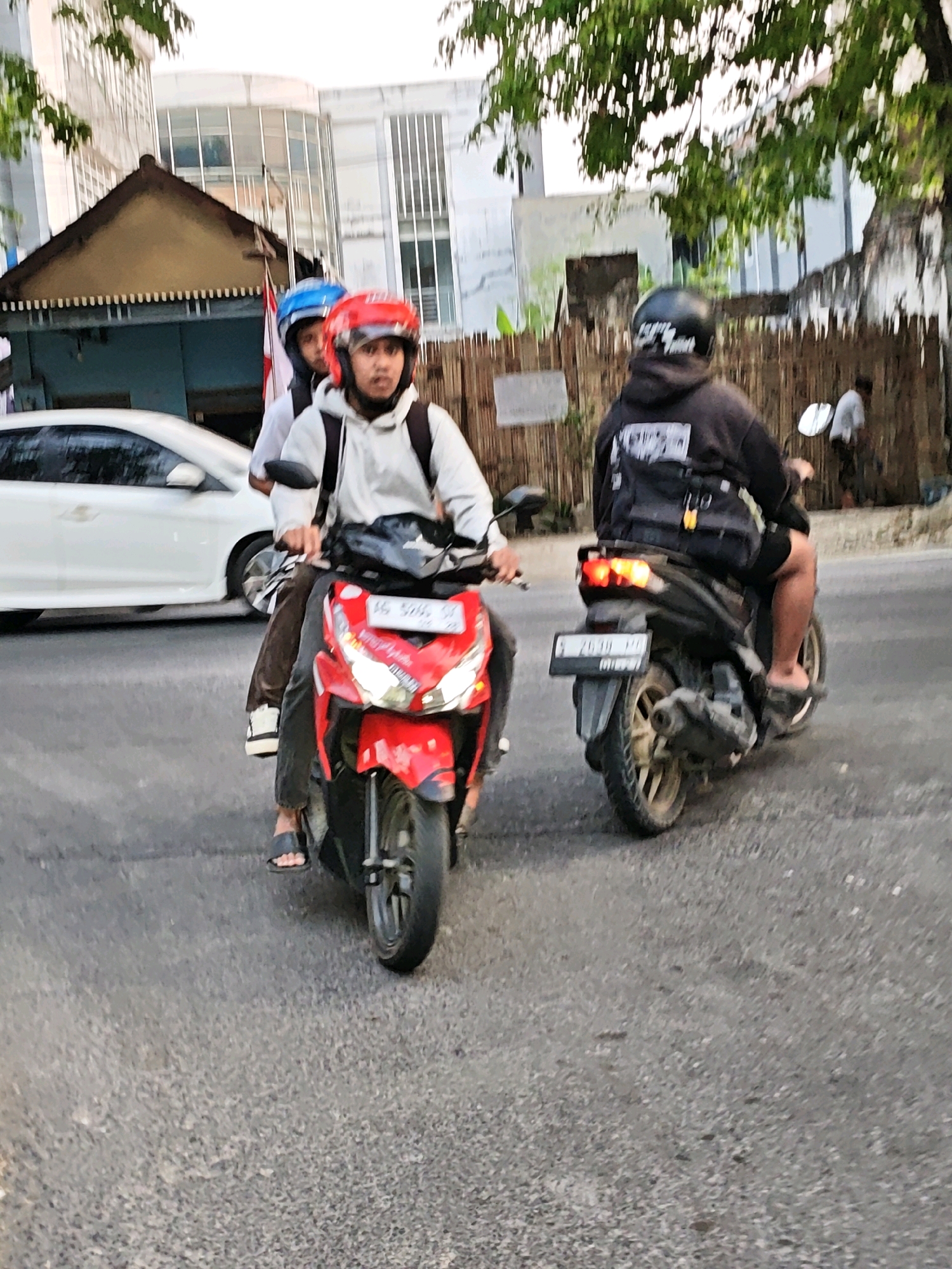 In the city of Kediri, Indonesia, three individuals are captured on motorcycles on a street. The foreground shows a red motorcycle with two occupants wearing helmets. The rider in the front wears a red helmet, and the passenger behind him wears a blue helmet. Both appear to be looking forward. To the right, another motorcycle is visible with a single rider wearing a black helmet, also facing forward. This rider is wearing a black hoodie and black shorts. The motorcycles are on an asphalt road with some debris. In the background, a white car is parked on the left side of the road. Buildings with architectural features like a triangular roof and windows are visible behind the motorcycles and car. Green foliage from trees is present in the upper portion of the image. A wooden fence runs along the right side of the scene. The lighting suggests it might be late afternoon or early morning.