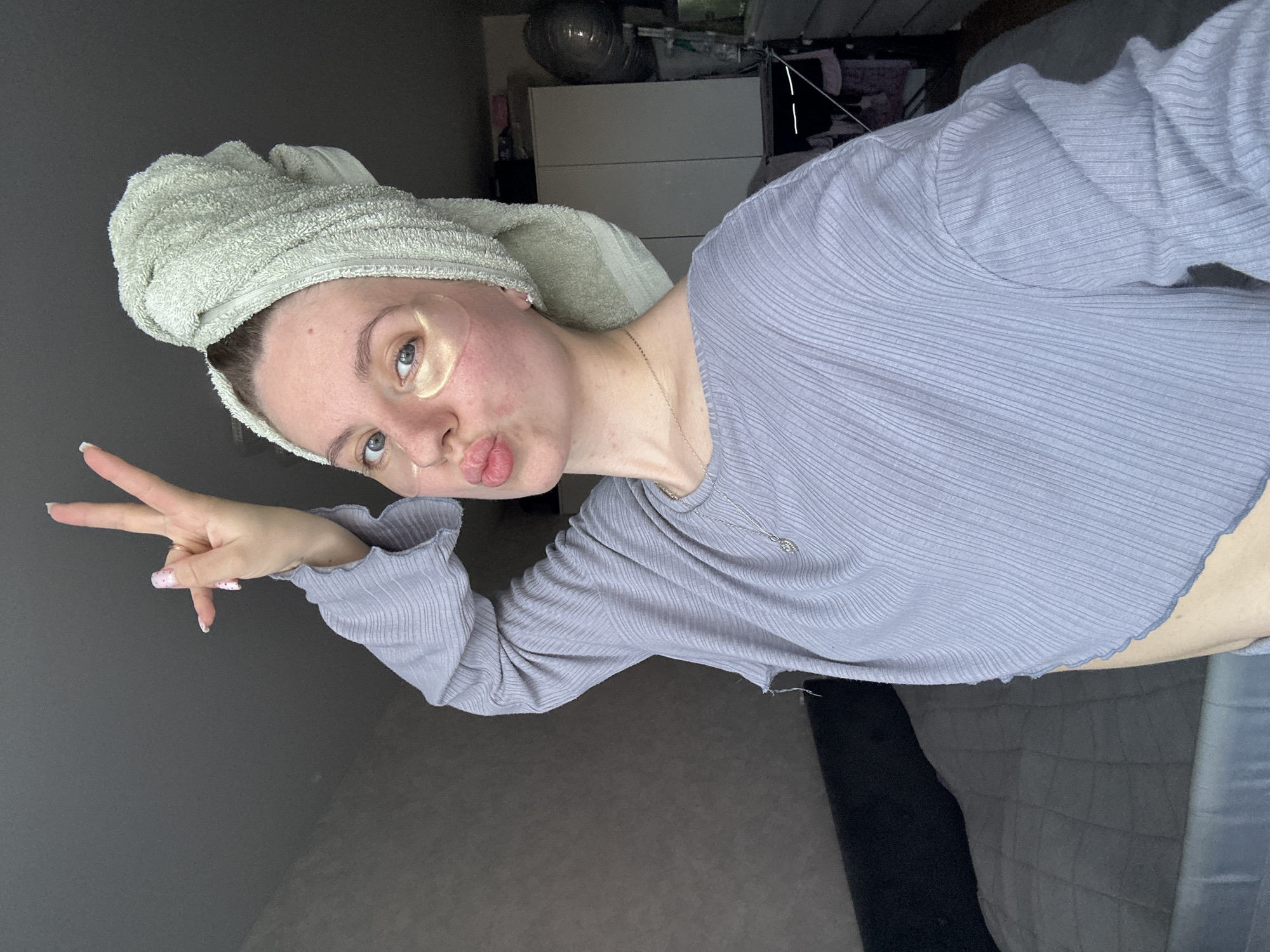 A woman is posing for a selfie. She has a towel wrapped around her head and gold under-eye patches on. She is wearing a long-sleeved shirt and making a peace sign with her left hand. The location appears to be an indoor setting. The location of the city of Klin, Russia is not discernible from the image.