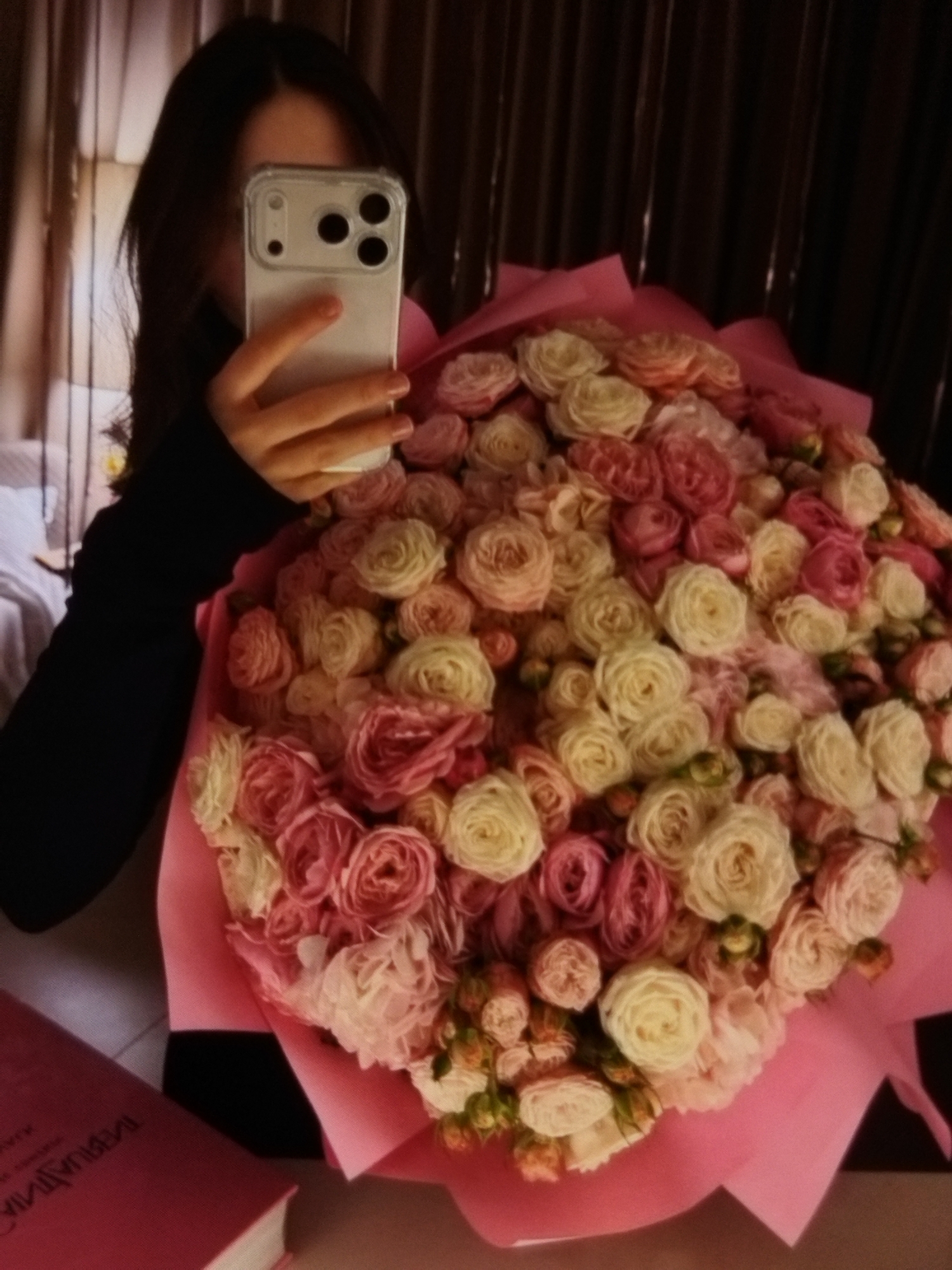 In Beșalma, Moldova, a person is taking a selfie while holding a large bouquet of pink and white roses. The person's face is obscured by a smartphone. The bouquet is wrapped in pink paper and is the central focus of the image. A pink book is visible in the lower left corner, with text on its cover. The background appears to be an indoor setting, possibly a bedroom, with curtains visible.