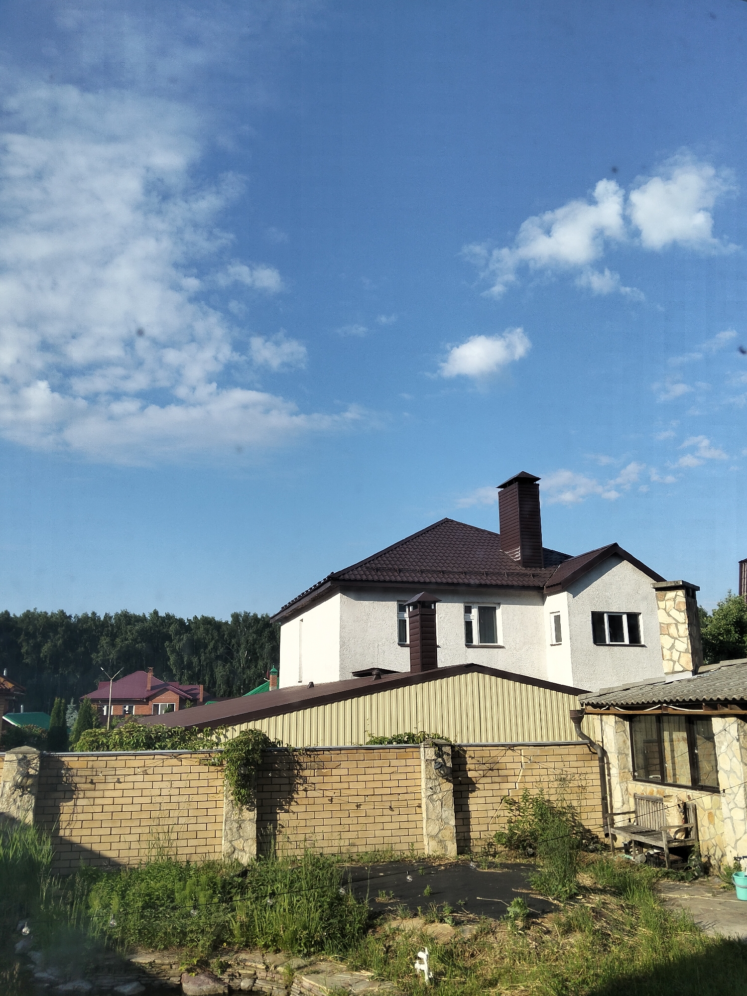 The image shows a suburban scene under a clear blue sky with scattered white clouds. In the foreground, a low brick wall with stone pillars separates the viewer's perspective from the property behind it. Overgrown green grass and weeds cover the ground in front of and on top of the wall.
Behind the wall, a two-story house with a dark brown tiled roof and white stucco exterior is visible. It has several windows with white frames and dark shutters or trim. A prominent dark brown chimney extends from the roof. Adjacent to this house is a single-story structure with a light yellow corrugated metal siding and a brown roof. Further back, a dense line of green trees forms a backdrop.
To the right of the main house, a smaller, single-story building with a stone facade and a grey, weathered roof is partially visible. A wooden bench sits in front of its windows. The overall impression is of a residential area on a sunny day.