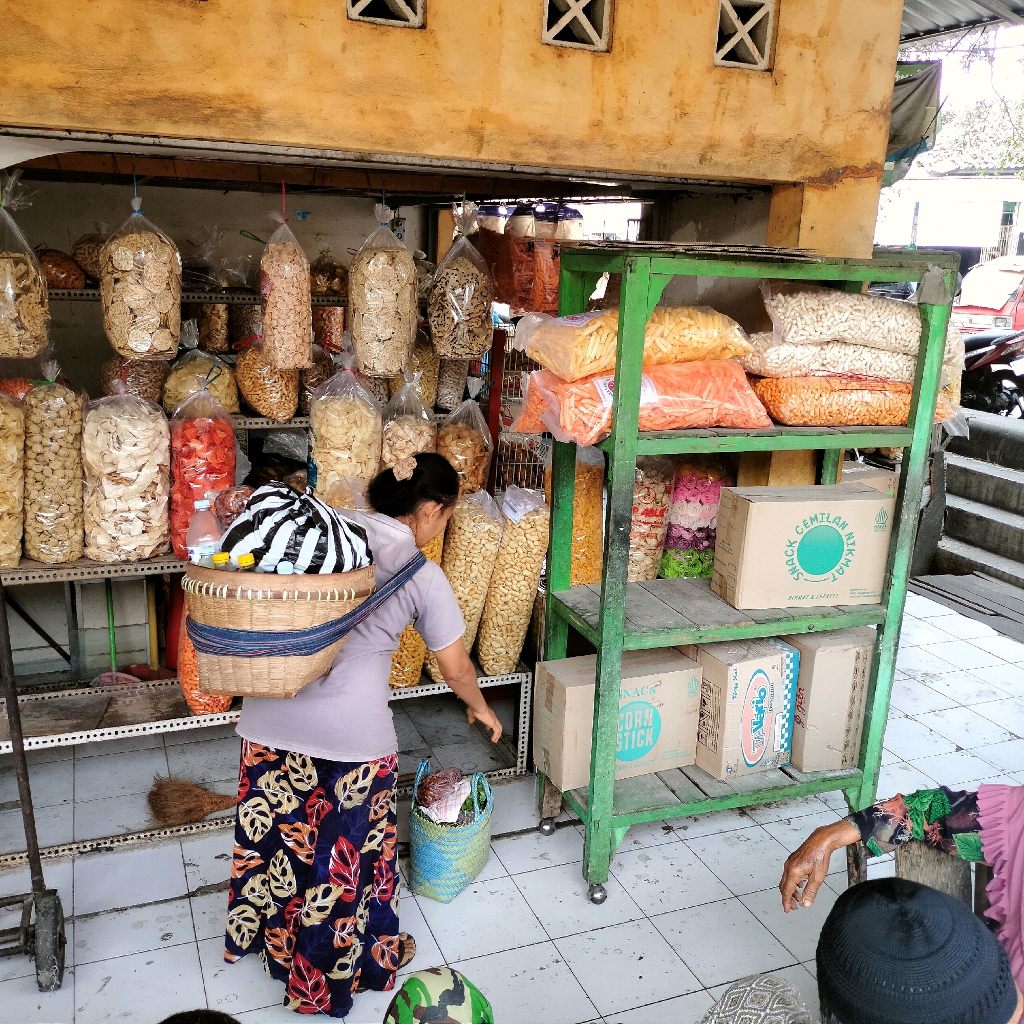 In an outdoor market stall in Wonosari, Indonesia, a woman wearing a patterned skirt and a grey shirt carries a woven basket on her back. The basket contains a striped bag, two bottles, and other items. She is reaching down towards a blue and green woven basket on the tiled floor, which holds some food items. The stall is stocked with various bagged snacks, including chips and puffed corn snacks, displayed on metal shelves and hanging from the ceiling. To the right, a green metal shelving unit holds boxes of snacks, with a staircase and part of a red vehicle visible in the background. The stall has an aged, yellow-painted wall above it, with ventilation openings.
