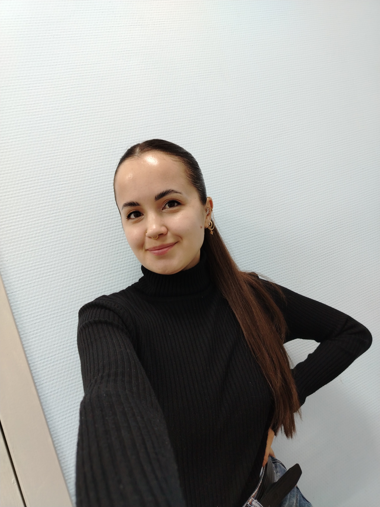 A young woman is posing indoors. She has long, straight, dark brown hair pulled back from her face into a low ponytail. She is wearing a black turtleneck sweater and jeans. Her skin is fair, and she has a small nose ring and multiple earrings in her left ear. She is smiling slightly. The background is a textured, light blue or white wall, which is probably located in the city of Chaikovsky, Russia.