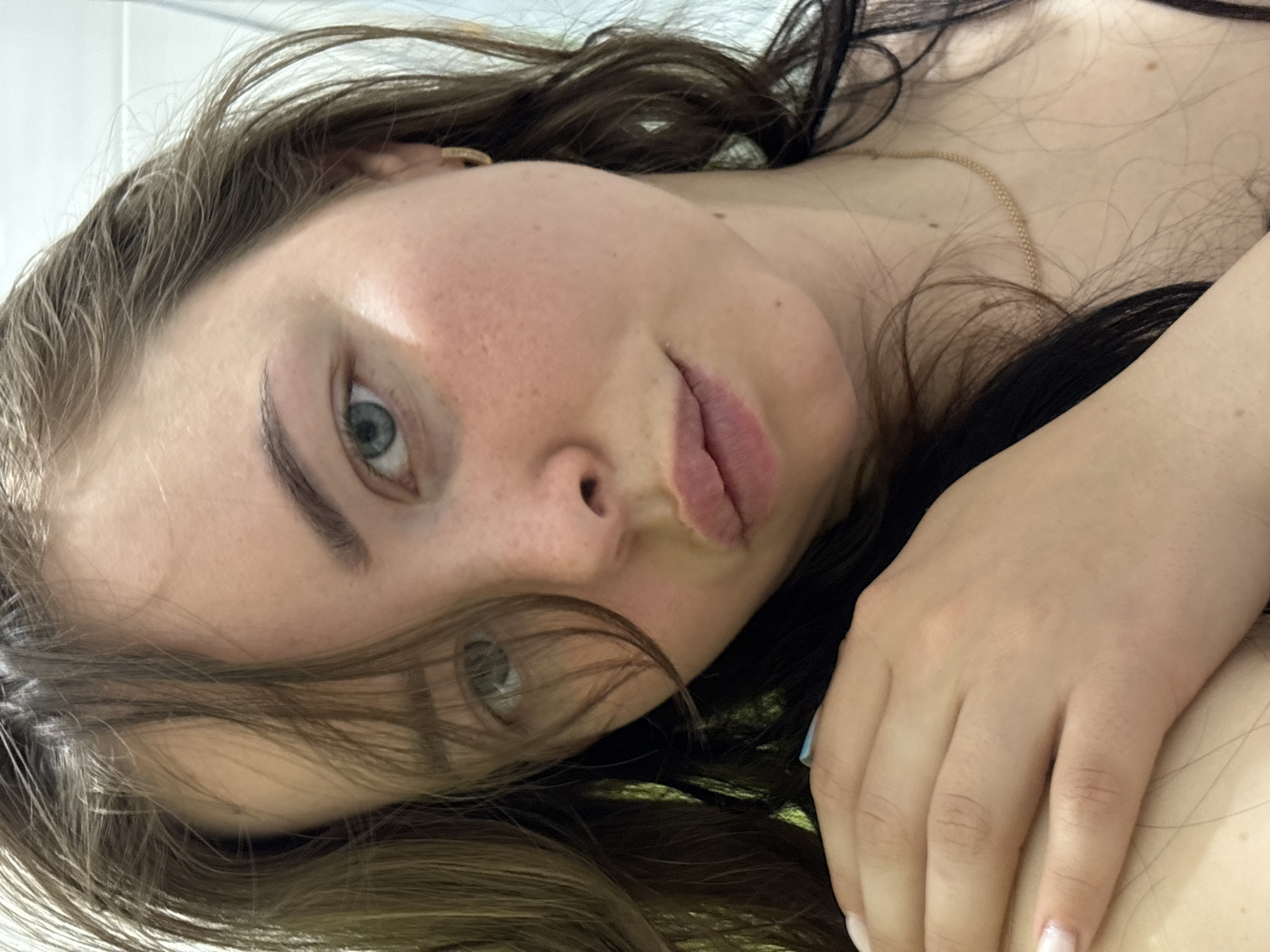 A close-up, high-angle portrait features a young person, likely female, lying down and looking directly into the camera. The image is oriented diagonally, with the person's head tilted.
The subject has fair skin, blue-green eyes, and dark eyebrows. Their lips are slightly parted. Long, wavy brown hair frames the face and spreads around the head and upper body. A thin, gold-colored chain necklace is visible on the chest, intertwined with strands of hair. Part of a hand rests on the shoulder/upper chest area, revealing a light-colored manicure on the fingernails, and a hint of light blue on one nail.
The background is simple and light-colored, suggesting an indoor setting, possibly a bed or sofa. The lighting is soft, highlighting facial features and skin texture. The overall mood is relaxed and casual. There is no visible text in the image. This scene takes place in Izhevsk, Russia.