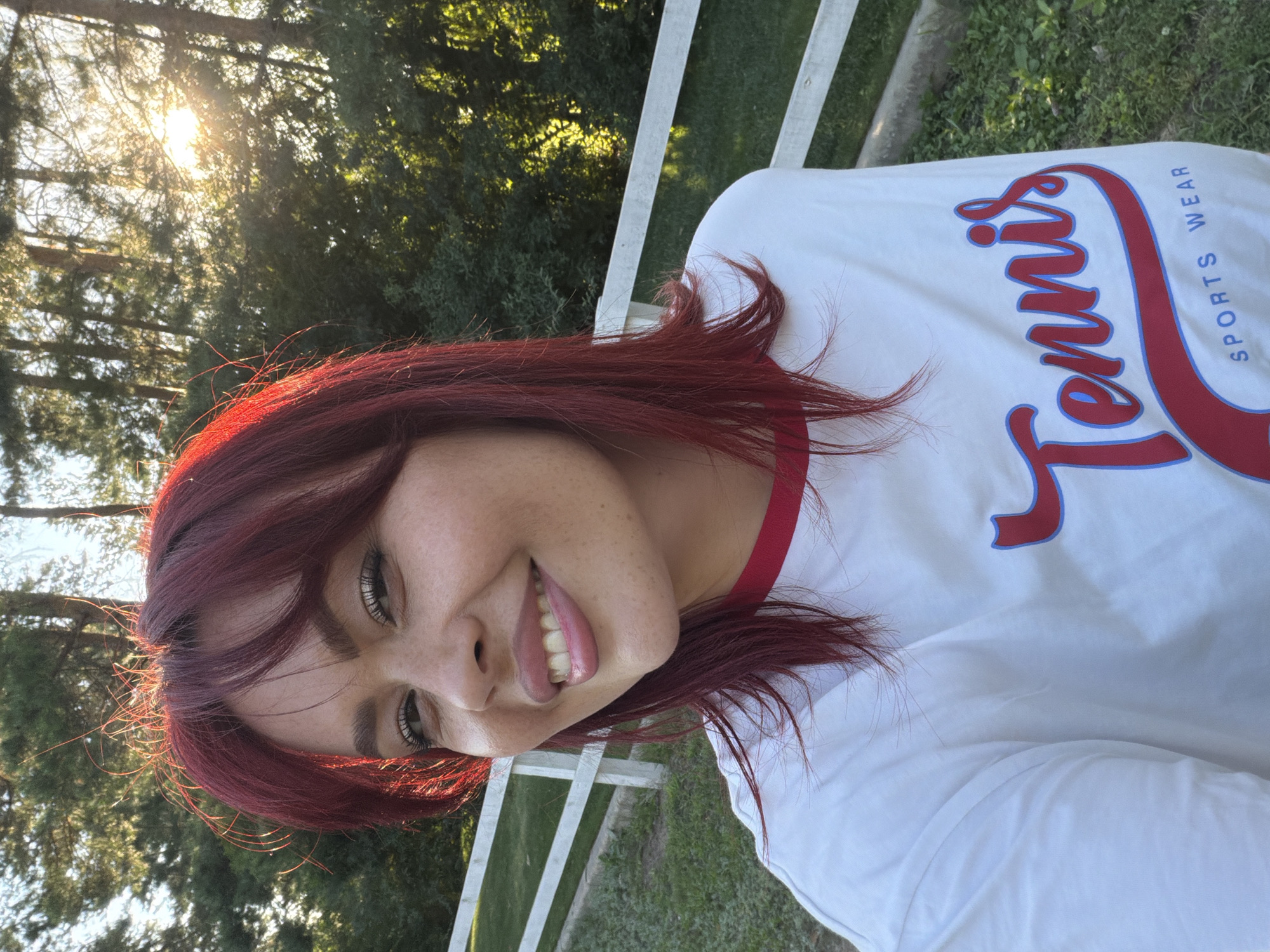 A young woman with vibrant red hair and fair skin, adorned with freckles, smiles while taking a selfie outdoors. She wears a white t-shirt with the word "Tennis" in red and blue lettering across the chest. The background features a white wooden fence, lush green grass, and trees bathed in warm sunlight. The scene appears to be a peaceful outdoor setting, possibly in a park or rural area, given the natural environment. There is no indication of the city of Узюково, Russia in the image.