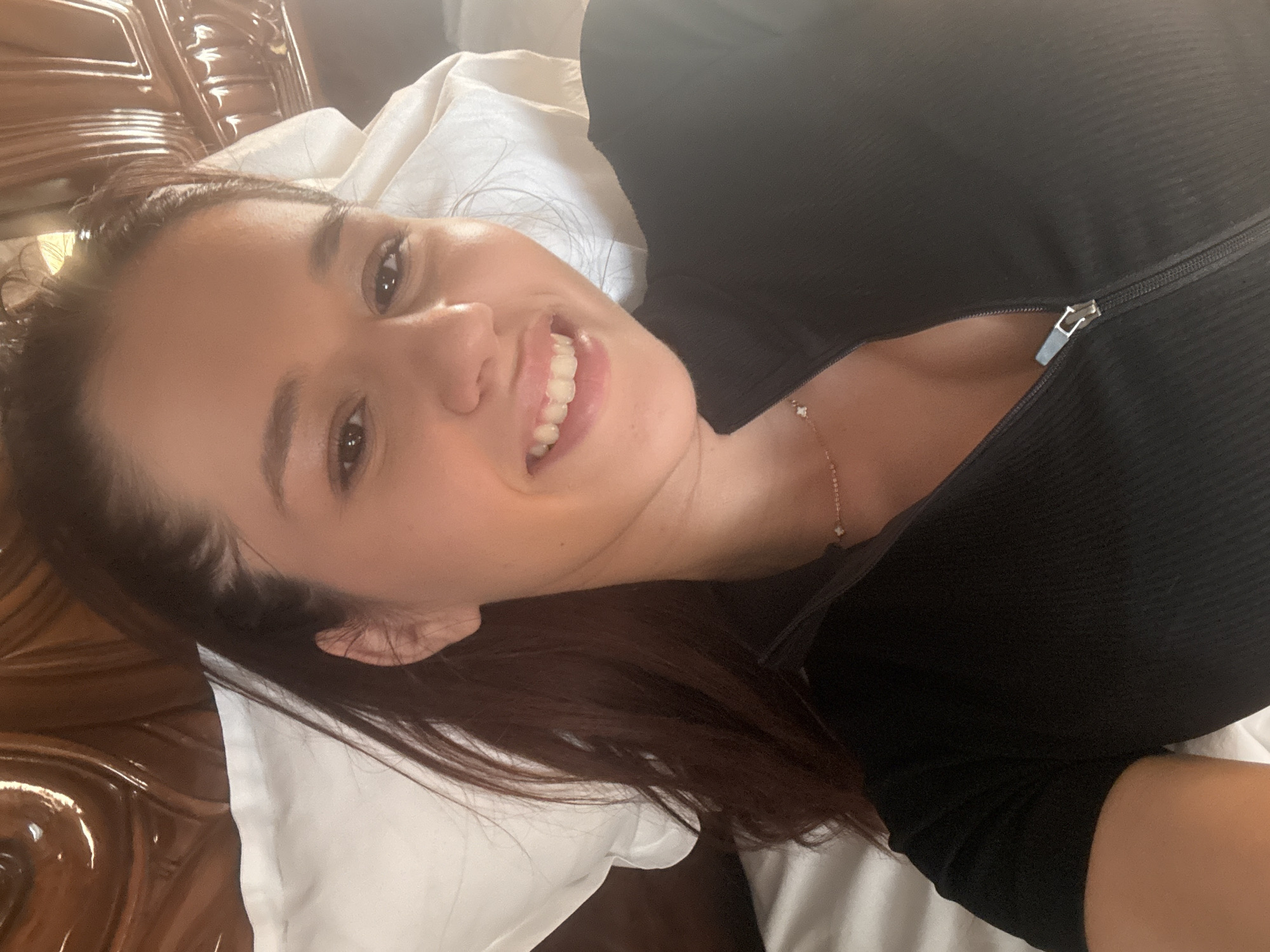 The image shows a woman lying on a bed, possibly indoors. She is smiling and looking towards the camera. She is wearing a black ribbed top with a zipper at the neck. A delicate necklace is visible on her neck. The background includes a white pillow and a wooden headboard. The location is not evident from the image; the assumption of Novo Zhelezare, Bulgaria, cannot be confirmed visually.