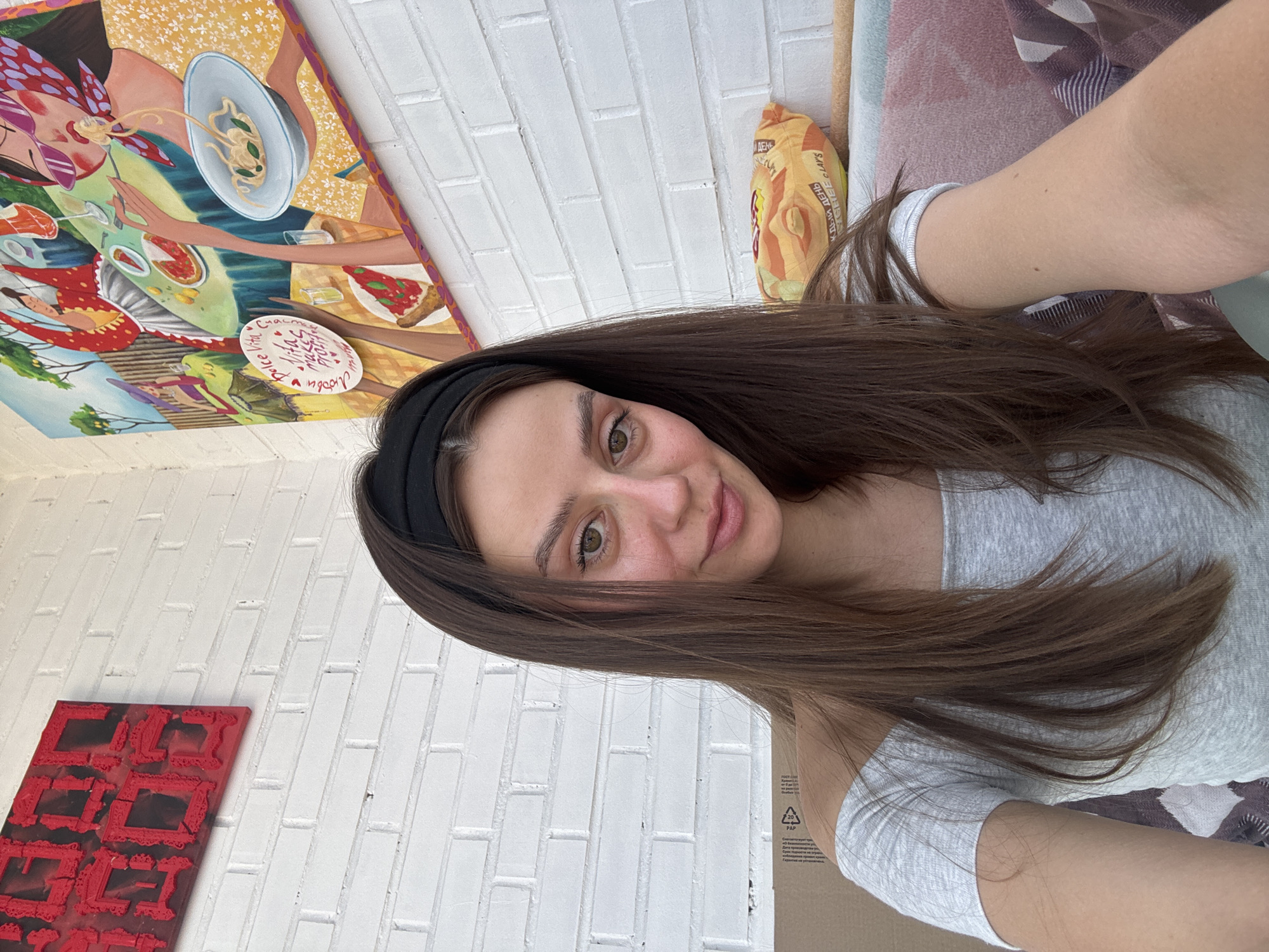 A young woman, appearing to be in her early twenties, is positioned centrally in a self-portrait, smiling slightly. She has long, straight, dark brown hair, a black headband, and light-colored eyes. Her skin is fair, with visible redness on her cheeks. She wears a light grey off-the-shoulder short-sleeved top, and small moles are visible on her right forearm. The background features a white brick wall.
To the upper left, a rectangular art piece with a red background and dark, ornate, framed shapes is mounted. To the upper right, a large, colorful painting depicts an outdoor dining scene with multiple women. One prominent figure on the right is eating spaghetti, wearing sunglasses and a patterned headscarf. Another figure holds a round sign with English text "Dolce Vita Ciao Bella" and "Vita makes profit," alongside Cyrillic text including "Любови" (of love) and "Каждыи день" (every day). A pizza slice is also depicted on a table in the painting.
Behind the woman's left shoulder, a brown cardboard box with visible packaging symbols is present. Behind her right shoulder, on a surface below the painting, a yellow-orange bag of Lay's potato chips is visible, displaying the Cyrillic inscription "КАЖДЫИ ДЕНЬ С LAY'S" (Every day with Lay's). Beneath the chip bag, a section of patterned fabric, in shades of pink, purple, and brown, is partially visible. The presence of Cyrillic text on the painting and the chip bag indicates a location within a Russian-speaking country. However, there are no specific visual cues within the image to definitively identify the city as Krasnoyarsk, Russia.