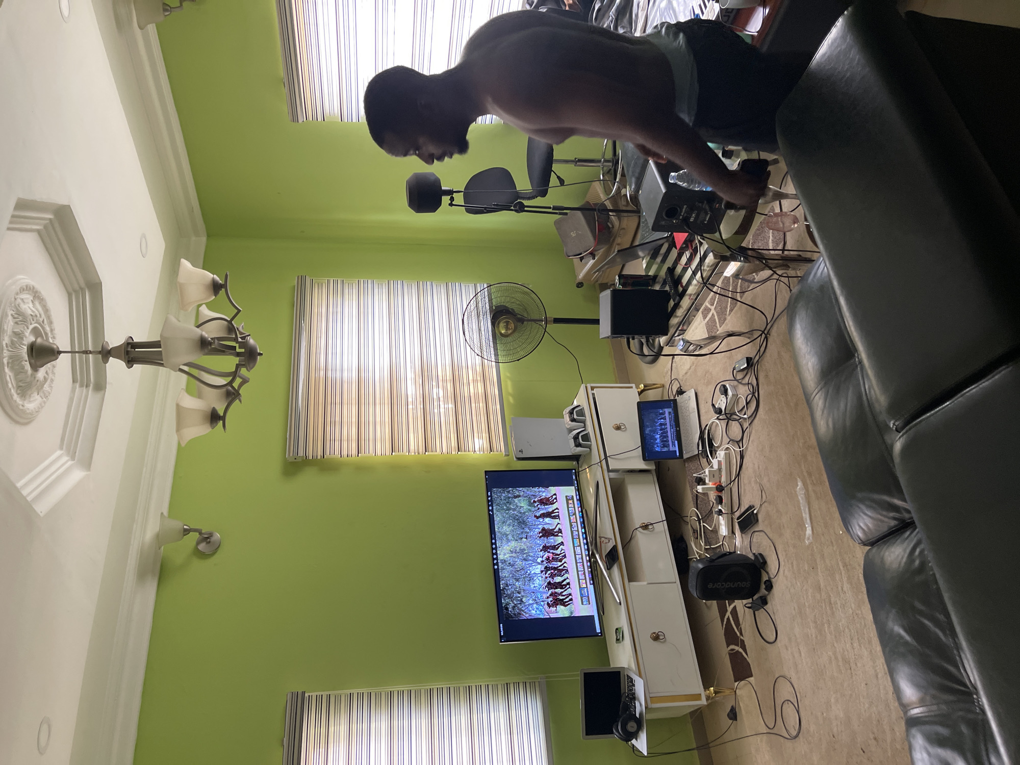 A man is doing push-ups in a room. The room has green walls, a chandelier, and two windows with blinds. A television is mounted on the wall. A desk fan is positioned in front of one of the windows. A black sofa is partially visible in the right side of the image. There is electronic equipment, including a computer and monitor, on a white cabinet. The location is in Abuja, Nigeria.