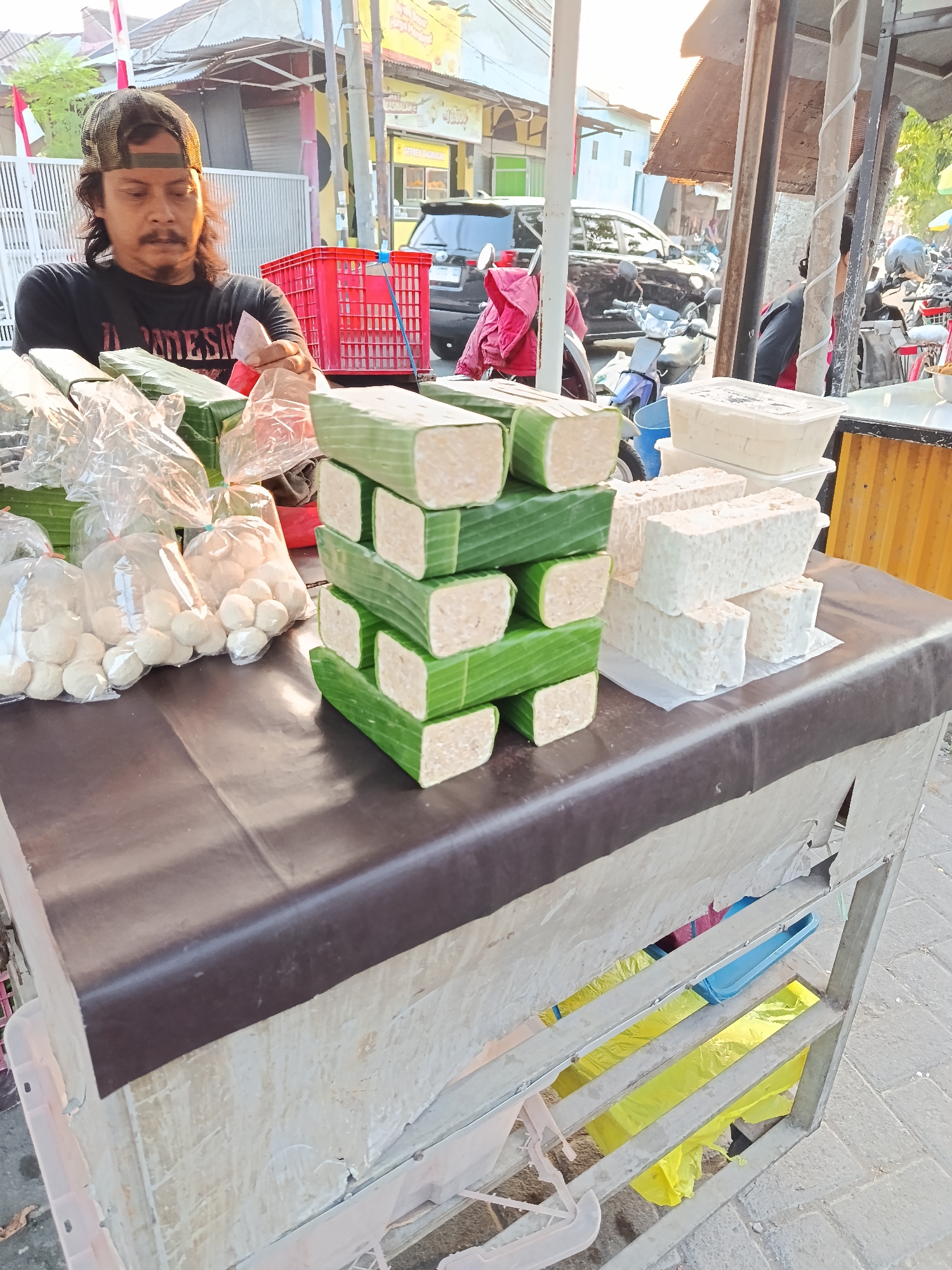A man with a mustache and dark hair wearing a black t-shirt and a cap stands behind a table laden with food items. He is holding a piece of food in his right hand, which is wrapped in plastic. In front of him are stacks of food wrapped in green banana leaves and white packaging. Behind him, a red crate is visible, along with a dark-colored car and motorcycles. The scene appears to be an outdoor market stall in Keboansikep, Indonesia, with buildings and a street visible in the background. The lighting suggests it is either early morning or late afternoon.
