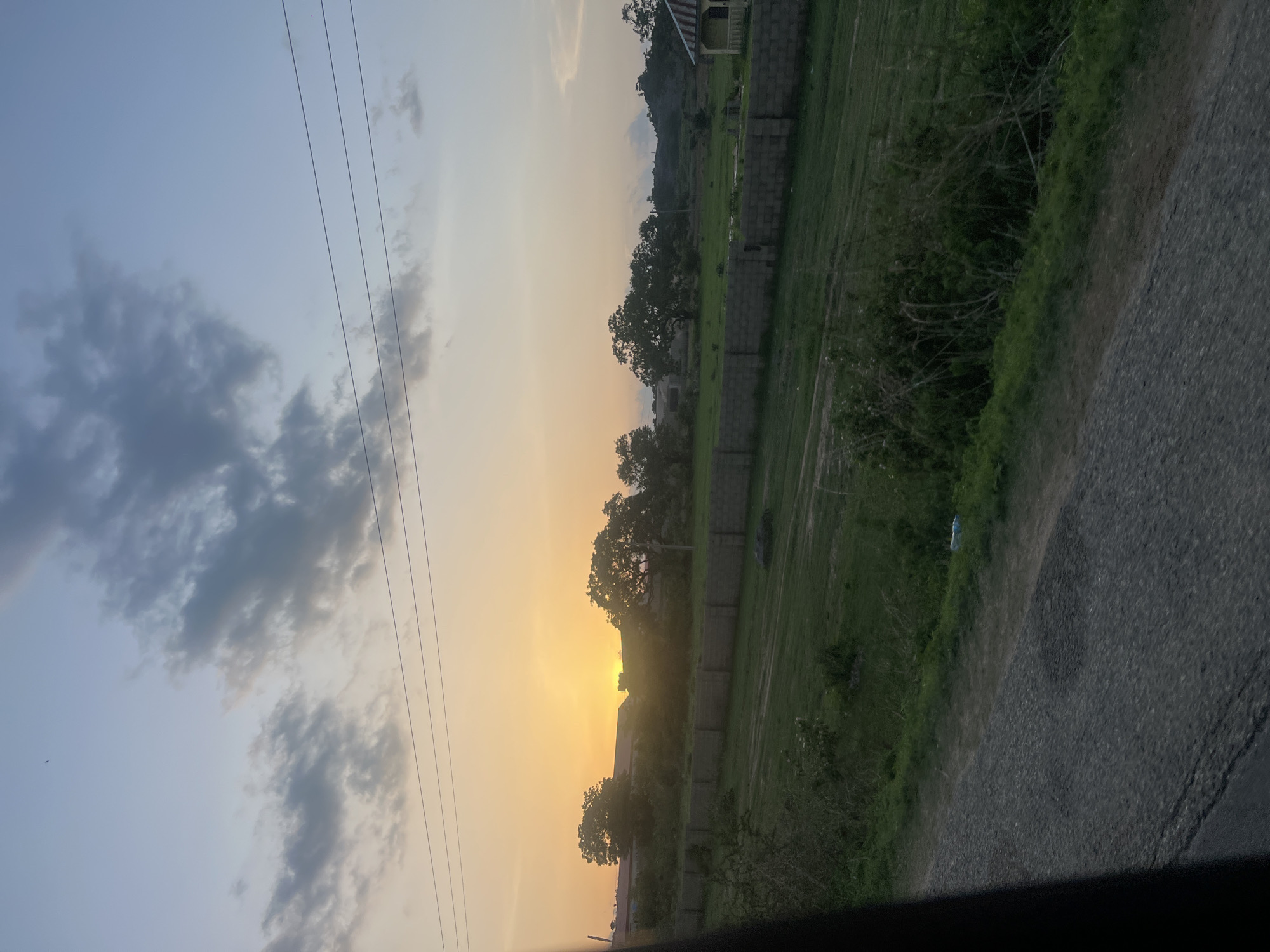 This photograph, taken in Rinjim Gani, Nigeria, depicts a sunset over a semi-rural landscape. The sun, a bright golden disc, is positioned low on the horizon on the left side of the frame, partially obscured by the silhouettes of structures and dense foliage. The sky exhibits a gradient, transitioning from a warm orange and yellow near the horizon on the left to a pale blue in the upper right quadrant, with scattered grey clouds spanning the upper half. Three distinct power lines run horizontally across the upper mid-ground. A small, dark, unidentified object, possibly a bird, is visible in the upper central-left sky.
In the mid-ground, a long, grey, cinder block wall extends horizontally across the scene, separating a wide expanse of green grassy field from the area beyond. Behind the wall and field, several large trees are prominent, and the roofs of some buildings are visible. On the far right, a solitary, small, yellow-painted building with a dark, sloped roof is situated near the edge of the grassy area. Distant hills or elevated terrain are present in the background on the right.
The immediate foreground features a dark, textured surface, indicative of a gravel or unpaved road, which runs diagonally from the lower left towards the lower right. Adjacent to the road on the right is a strip of dense green vegetation and bushes. A dark, vertical element is visible along the far left edge of the image, suggesting the photograph was taken from within a vehicle. No people are present in the scene.