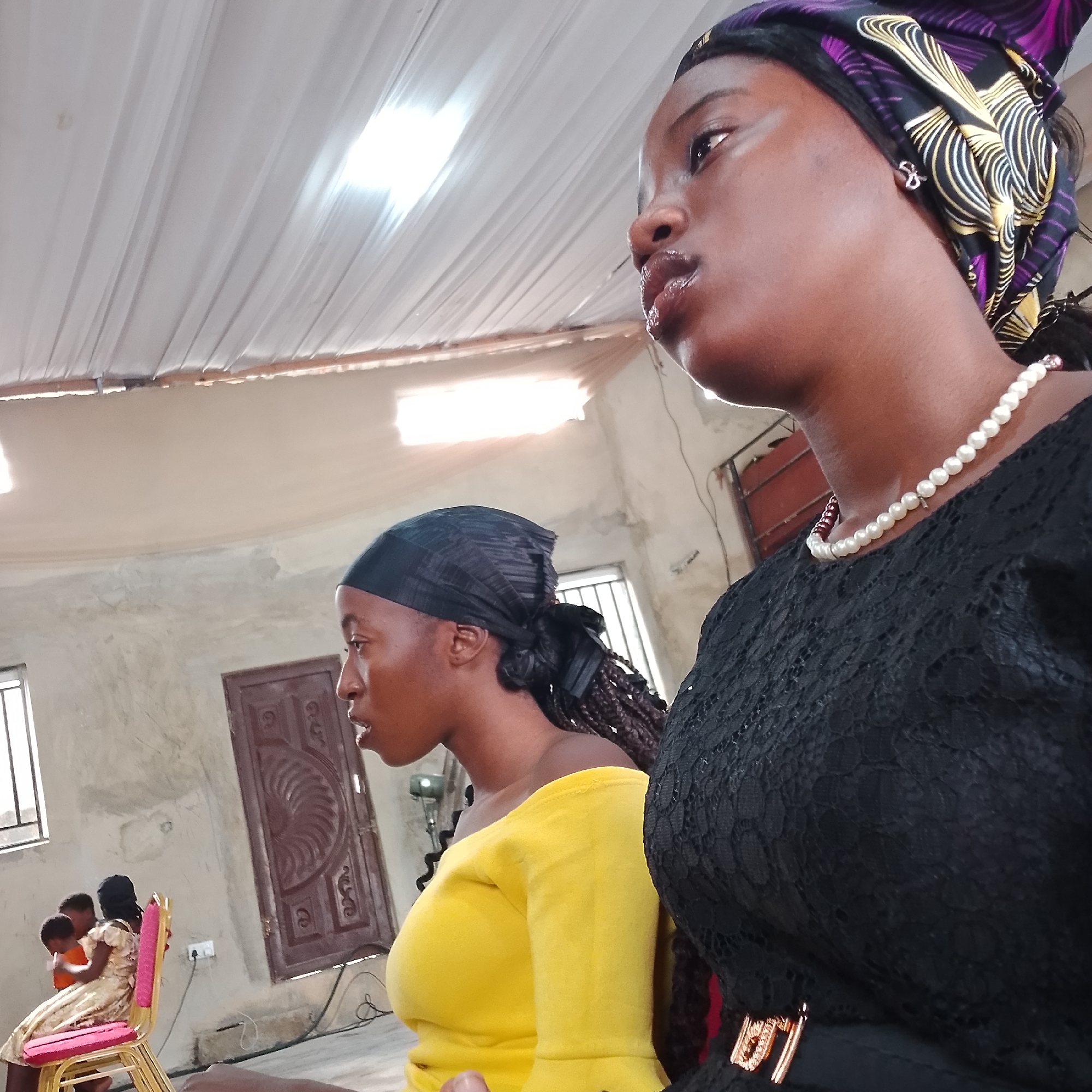 The image depicts an indoor gathering, possibly a religious service, taking place in a hall in Ado, Nigeria. Two young women are prominently featured in the foreground, facing towards the right of the frame. The woman on the right is wearing a black lace dress and a patterned headwrap with purple, yellow, and black motifs. She has a pearl necklace and is looking upwards with her mouth slightly open. The woman on the left is wearing a bright yellow off-the-shoulder top and a black headscarf. Her gaze is directed forward, and her mouth is also slightly open.
In the background, several other individuals, including children, are visible, seated on chairs. One child is wearing a light-colored dress. A wooden door with intricate carvings and a metal handle is visible on the wall behind the seated individuals. The room has plain, lightly colored walls and a ceiling covered with white fabric. Natural light appears to be entering the room from windows that are not fully visible, illuminating the space. The overall atmosphere suggests a community event or service.