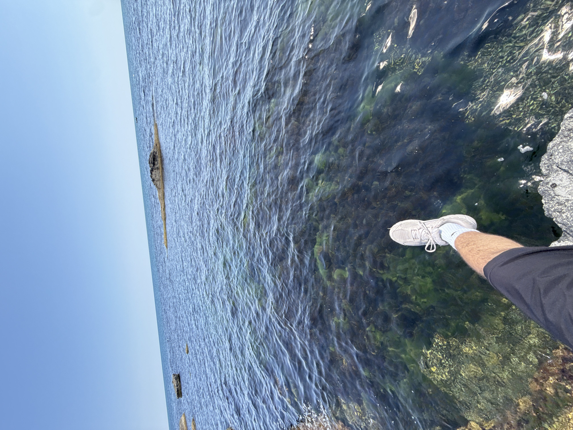 A person's legs and feet, clad in dark shorts, a light blue sock, and a white sneaker, are visible in the foreground, dangling over a rocky shoreline. The water is clear, revealing green seaweed and rocks beneath the surface. The sea extends to the horizon under a clear blue sky, with a few rocks visible in the distance. The scene is located at a rocky coast near Tsarevo, Bulgaria.