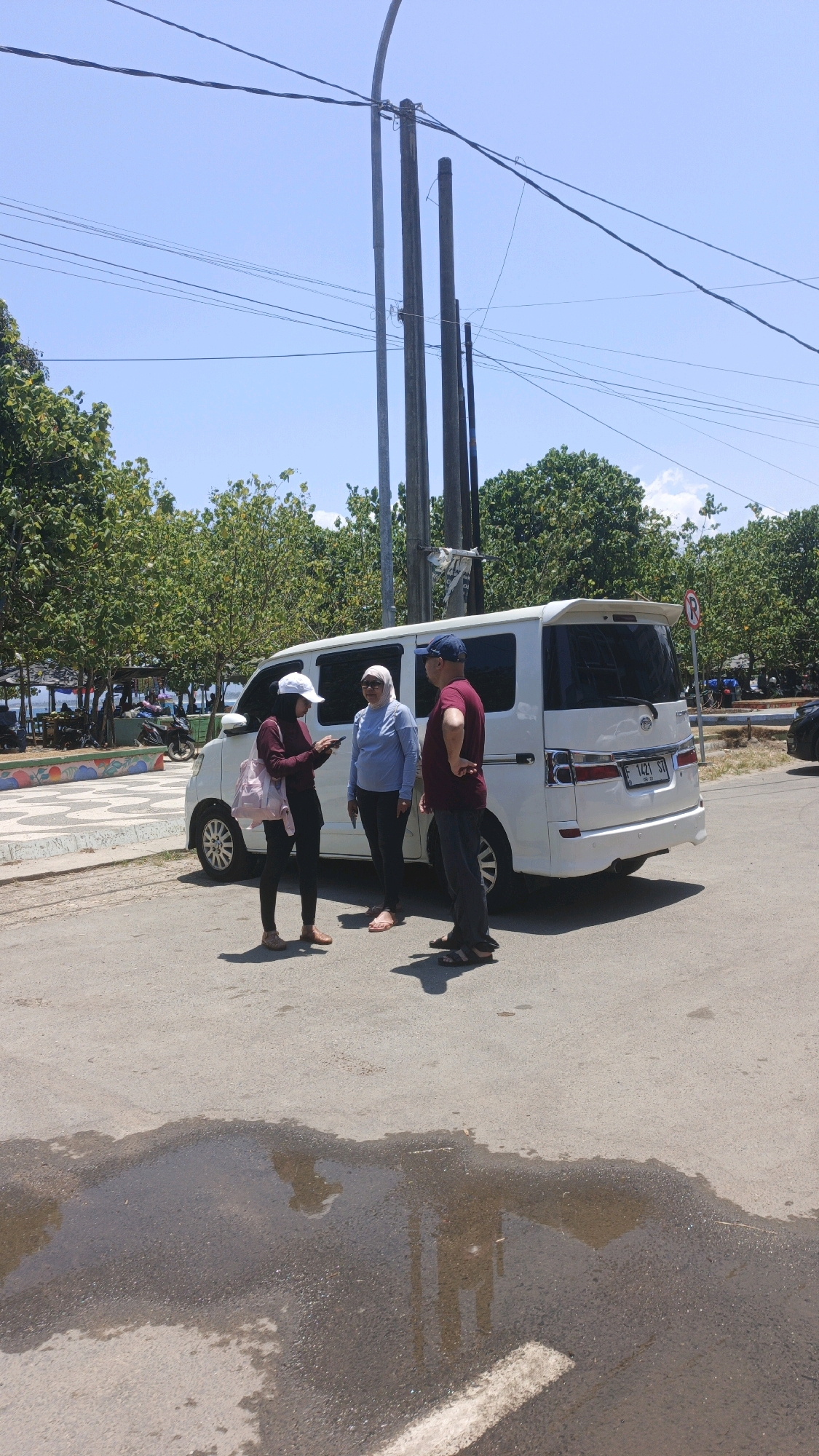 Three individuals are standing next to a white minivan parked on an asphalt road in Desa Pagandaran, Indonesia. A woman wearing a maroon hijab and a white cap is looking at her phone, holding it with her right hand and her left arm extended. She is wearing a maroon jacket and black pants. To her right, a woman in a light blue hijab and a light blue long-sleeved shirt is facing the camera. To the right of the woman in blue, a man wearing a dark blue baseball cap, a maroon short-sleeved shirt, and dark pants is standing with his right hand on his hip, looking away from the camera. The minivan, which has a spoiler on the rear, has a license plate that reads "F 1421 ST". In the background, there are trees, a blue sky with scattered clouds, and overhead power lines supported by utility poles. Further back, a body of water can be seen with several small structures near the shore, and some parked motorcycles. A red and white circular sign with a diagonal red bar is visible on a pole to the right of the minivan. The asphalt road in the foreground has a large puddle reflecting the sky and the utility poles.