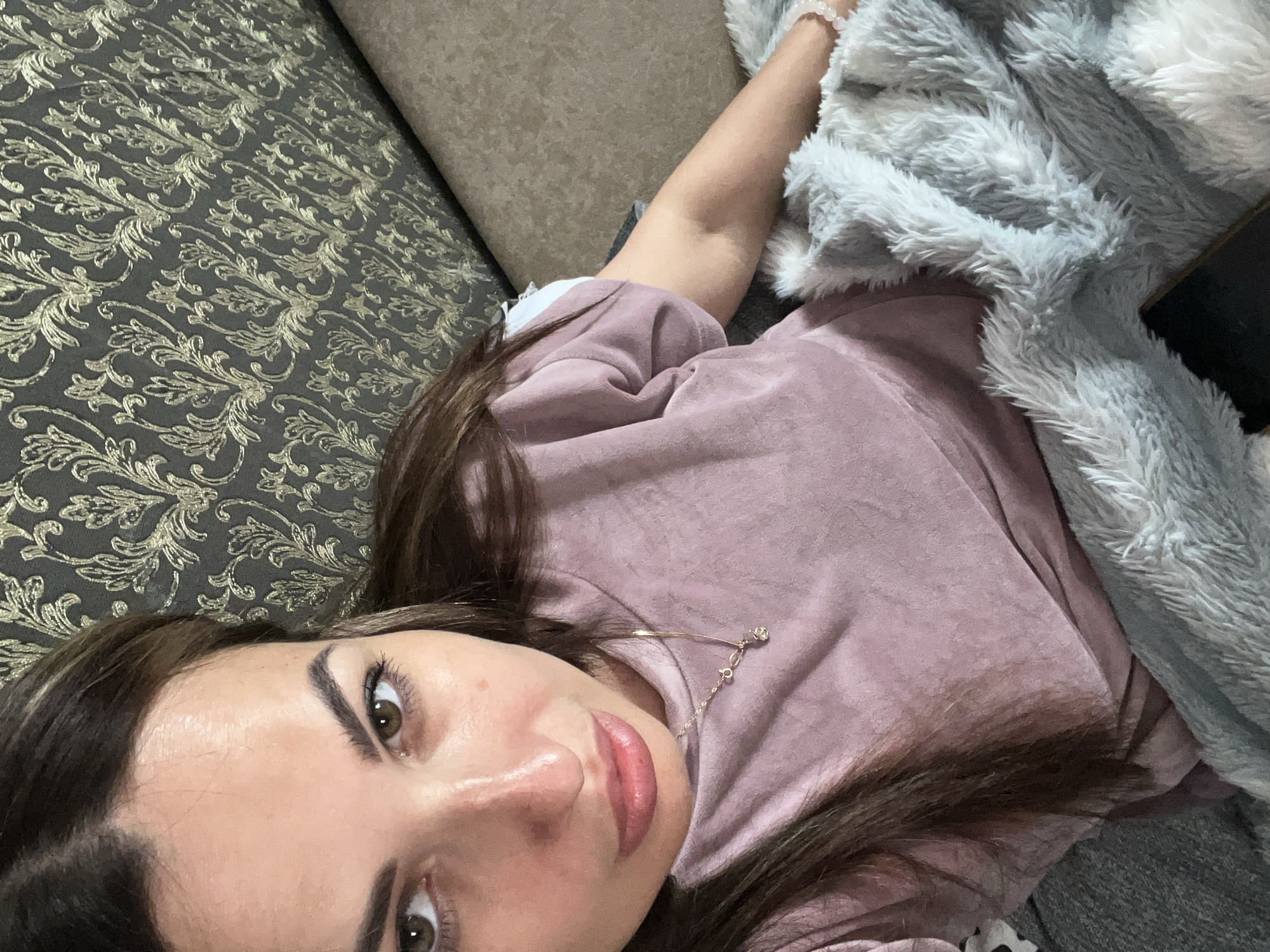 A young woman with long brown hair is lying on her side, covered by a fluffy grey blanket. She is wearing a pink shirt and has a necklace with a small charm. Her eyes are open and she is looking at the camera. The background appears to be a sofa with a patterned fabric. The lighting suggests it could be indoors, possibly in the evening or on a cloudy day, as the light is soft and diffused. The woman's expression is relaxed and content. There is no visible text in the image.