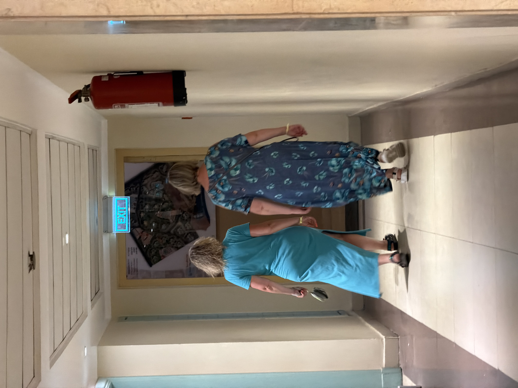 Two women are walking away from the camera down a hotel hallway. The woman on the left wears a bright turquoise maxi dress and sandals, carrying a small clutch. The woman on the right wears a dark blue floral maxi dress and sandals. An illuminated green "EXIT" sign is mounted on the ceiling ahead of them, and a red fire extinguisher is mounted on the wall to the right. The hallway has tiled floors and cream-colored walls, with ceiling tiles and recessed lighting. The image appears to have been taken in Sharm El Sheikh, Egypt.