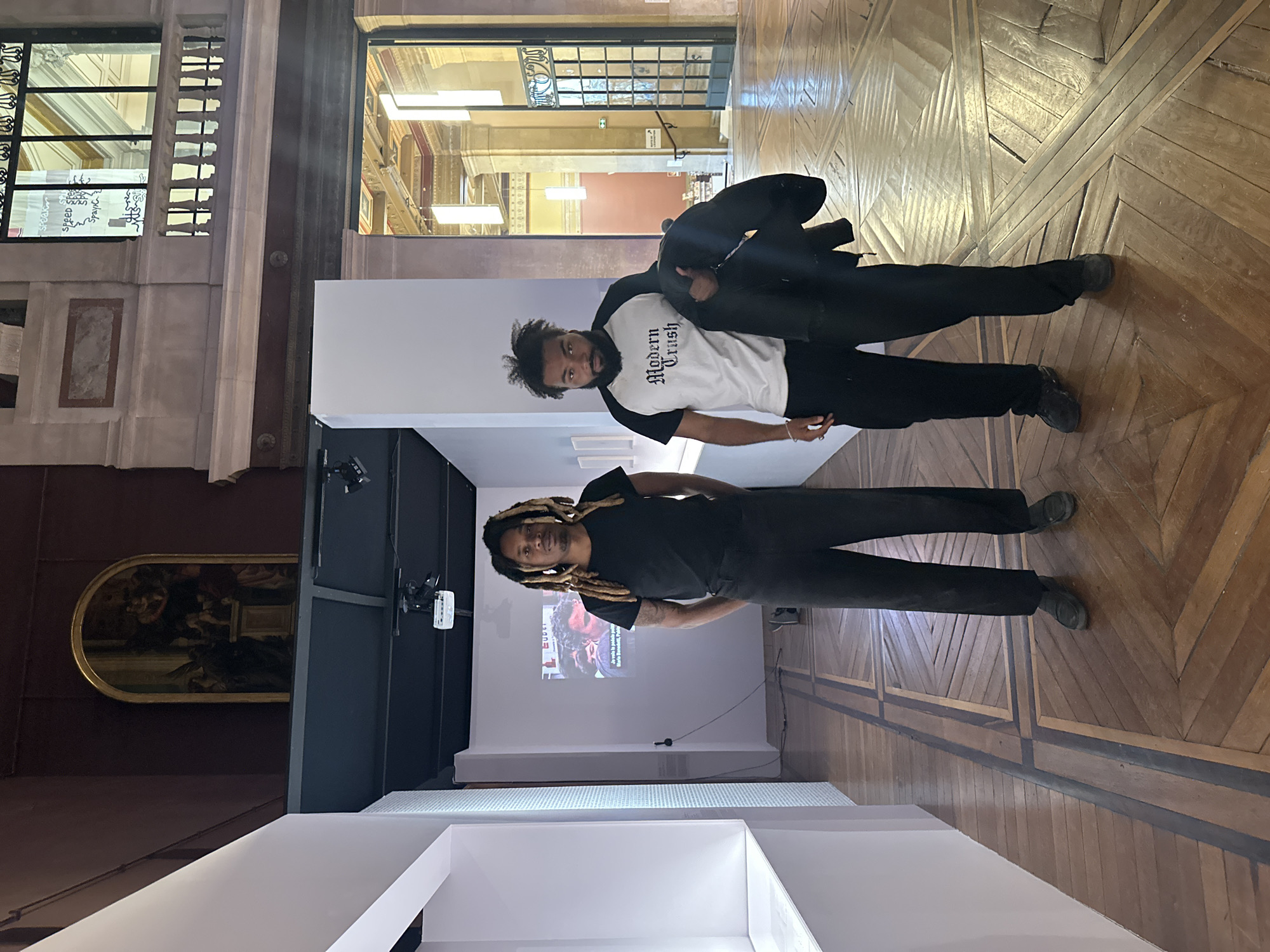 The image captures two men posing in what appears to be an exhibition space within a grand, possibly historic, building. The setting suggests an art gallery or museum in Paris, France, given the ornate architecture and wooden parquet flooring.
The man on the left has dreadlocks and is wearing a black t-shirt and dark trousers. He stands near a white projection screen displaying a close-up image of a person's face, possibly from a video or news report, with partially visible text like "Maria P..." and "Política." A projector is mounted on a dark structure behind him.
The man on the right, positioned further back, wears a white t-shirt with black sleeves and the text "Modern Crush" printed in a stylized font. He has a beard and short, dark curly hair, and is wearing dark trousers and black shoes. He holds a dark jacket draped over his shoulder. Both men are looking towards the camera with neutral to slightly smiling expressions.
The background features tall, elegant windows with decorative grilles, revealing interior architectural elements like arched doorways, columns, and modern lighting fixtures, suggesting another part of the building or an adjacent hall. In the upper left background, some white text, including "SPEEd" and "SPAINk," is visible on one of the windows. A large, framed painting with an arched top, depicting a classical scene, is visible on a dark wall behind the exhibition structures. The floor is made of richly patterned wooden parquet, adding to the historical ambiance. White modular display structures are also part of the exhibition setup.