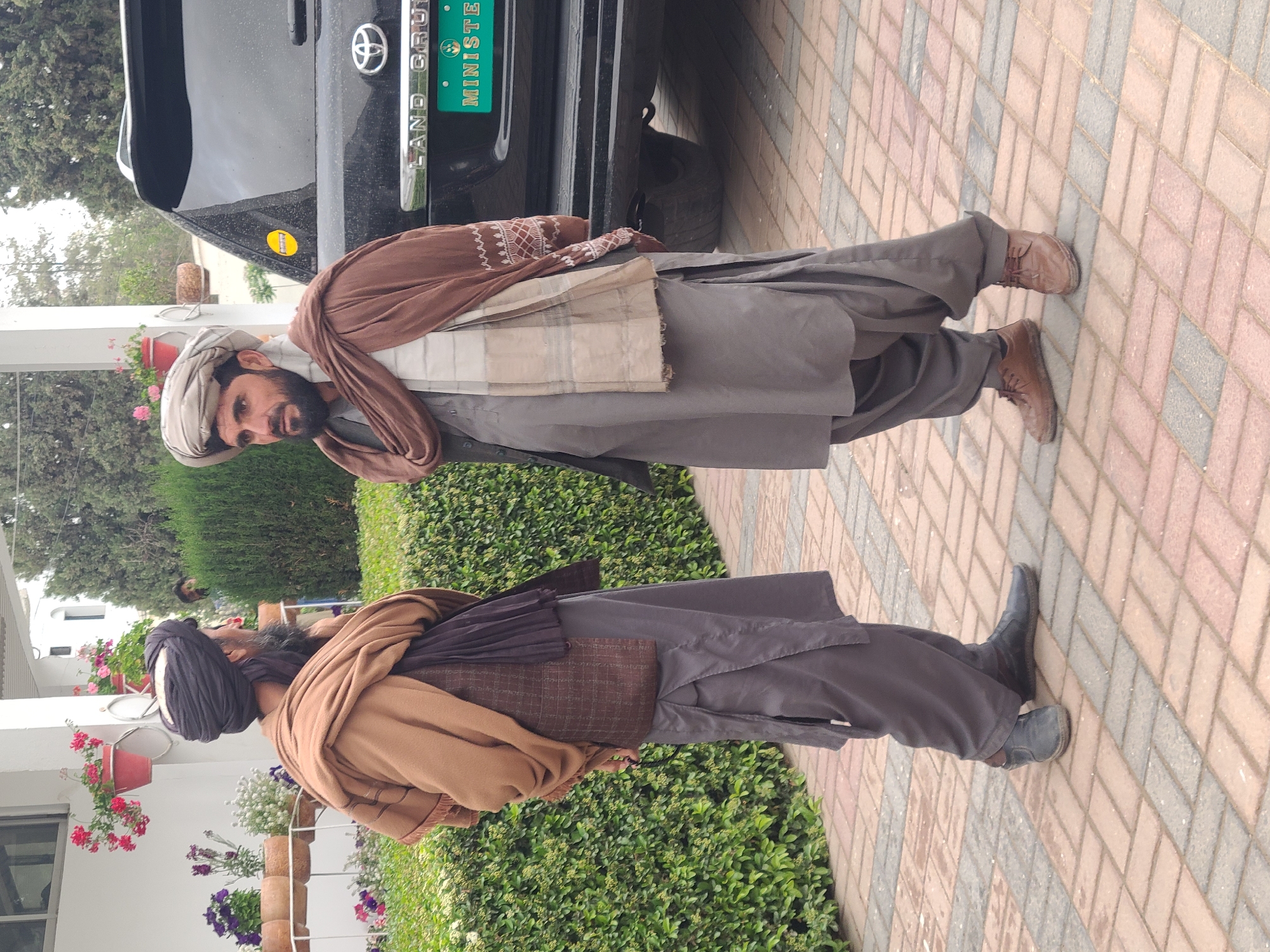 Two men stand in front of a black Land Cruiser in Quetta City Tehsil, Pakistan. The man on the right wears a light-colored turban, brown shawl, light gray traditional garb, and brown shoes. The man on the left wears a dark-colored turban, brown shawl, dark gray traditional garb, and black shoes. The vehicle behind them has a green license plate that reads "MINISTER." They stand on a brick sidewalk next to a hedge. A white building with flower pots is visible in the background.