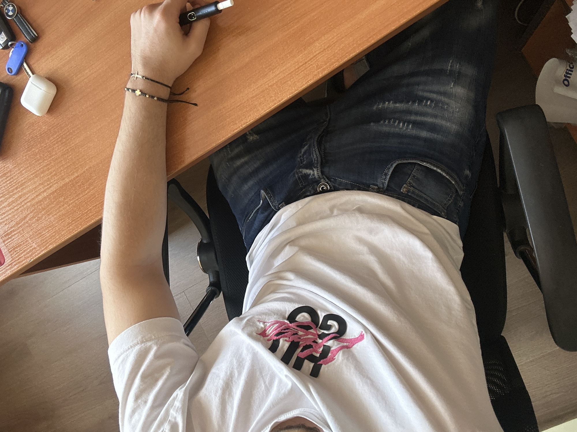 An aerial view looking down upon a person sitting at a desk. The person is wearing blue jeans and a white t-shirt. The desk is made of light brown wood. The person is holding a black electronic cigarette in their right hand. The person is wearing two thin bracelets. The environment includes keys, an airpods case, an office supplies box and a chair. The location is potentially a personal workspace. No location in the city of Plovdiv, Bulgaria is able to be identified.