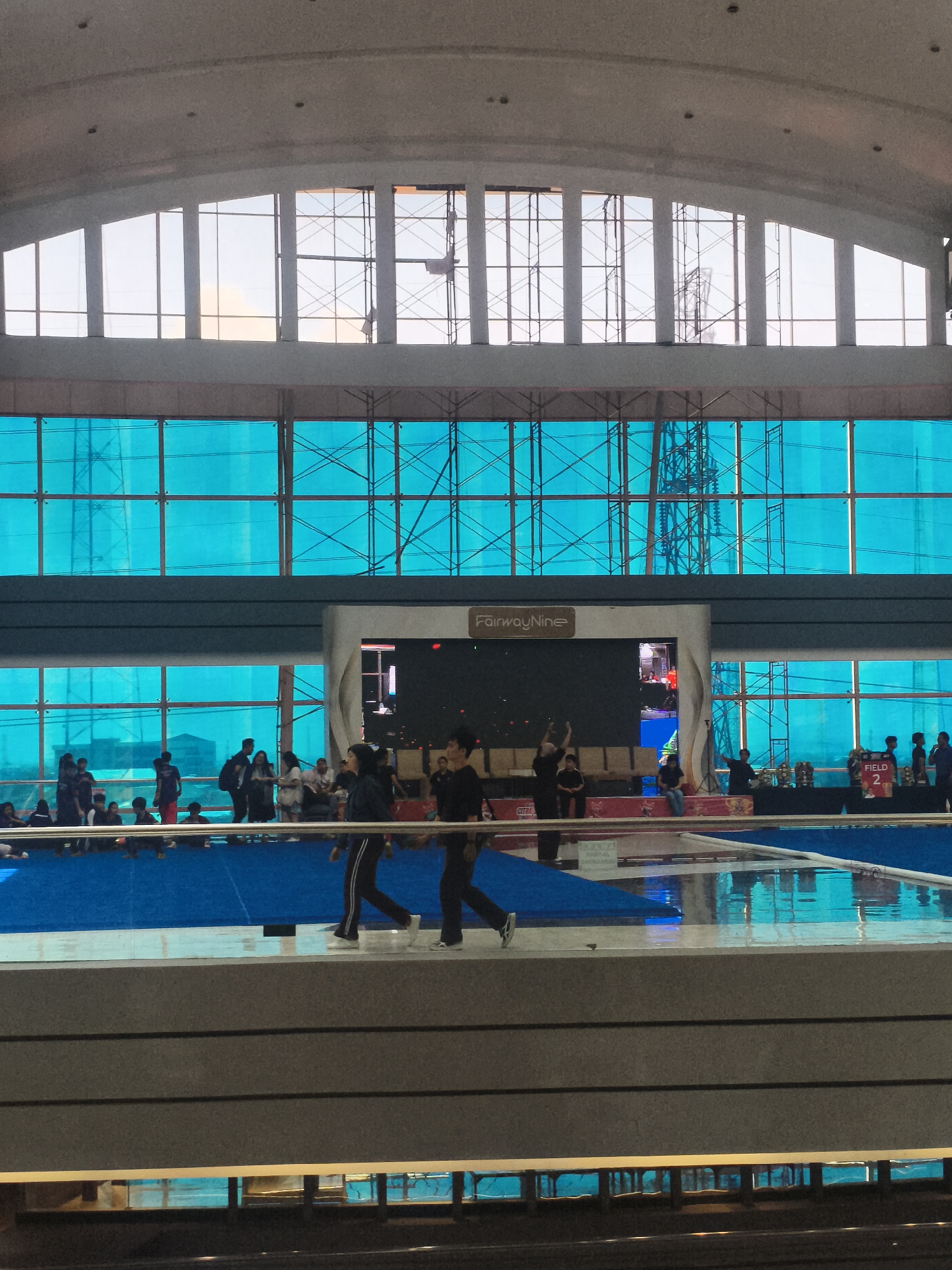 The image depicts the interior of the Juanda International Airport in Surabaya, Indonesia, specifically within the departure terminal. The scene captures a bustling atmosphere with numerous people present, suggesting ongoing travel activities. In the foreground, two individuals are walking across a polished floor, their attire indicating they are likely passengers or staff. Behind them, a blue matting is visible, possibly part of a waiting area or an event setup.
Further back, a large screen displays content, and a sign reading "FairwayNine" is prominently displayed, suggesting this area is managed or sponsored by this entity. Several individuals are seated or standing around this central area, some appearing to be spectators or participants in an event. Rows of empty chairs are also visible.
The background reveals a distinctive architectural feature: large windows with a turquoise tint, through which scaffolding and construction elements are visible, hinting at ongoing renovations or expansions within the airport. The overall impression is one of a dynamic and active airport environment, with a blend of everyday travel and potential special events taking place.