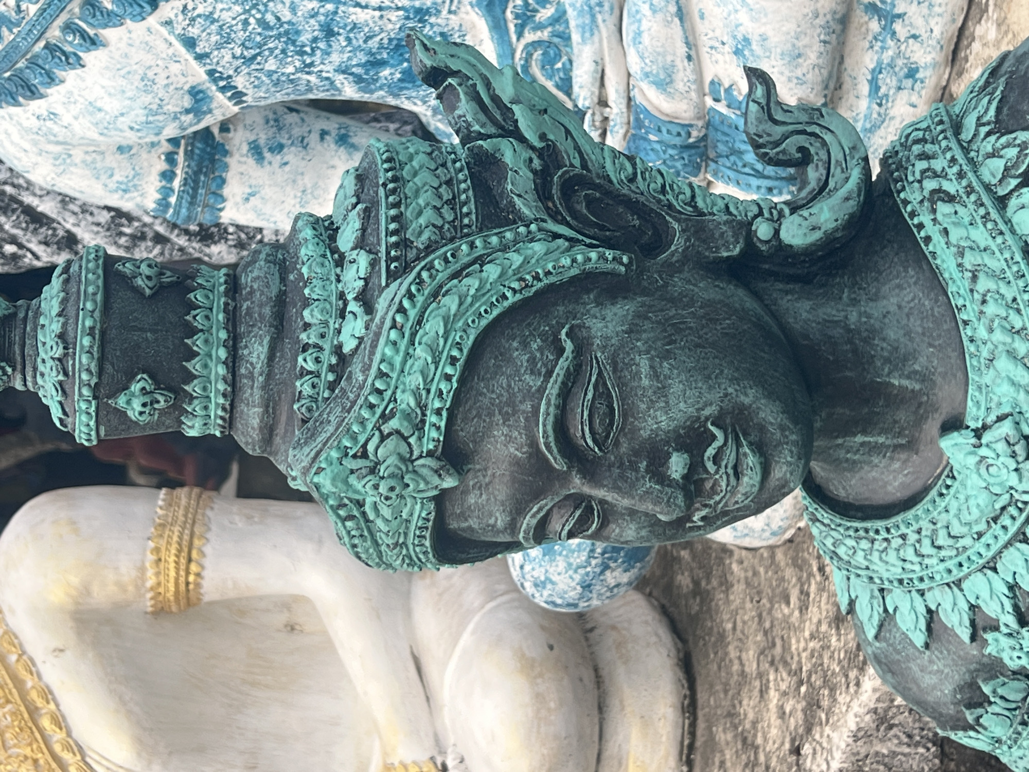 The image shows a close-up of a dark-toned, aged statue with teal-colored embellishments, possibly bronze with verdigris patina. The statue depicts a figure wearing an ornate headdress and collar. In the background, out of focus, are other statues, one appearing to be large and white with gold trim, and another in blue and white. A mottled, greyish ground is visible in the lower left. The setting appears to be an outdoor location in Thailand, possibly a temple or garden in the Ko Samui District.