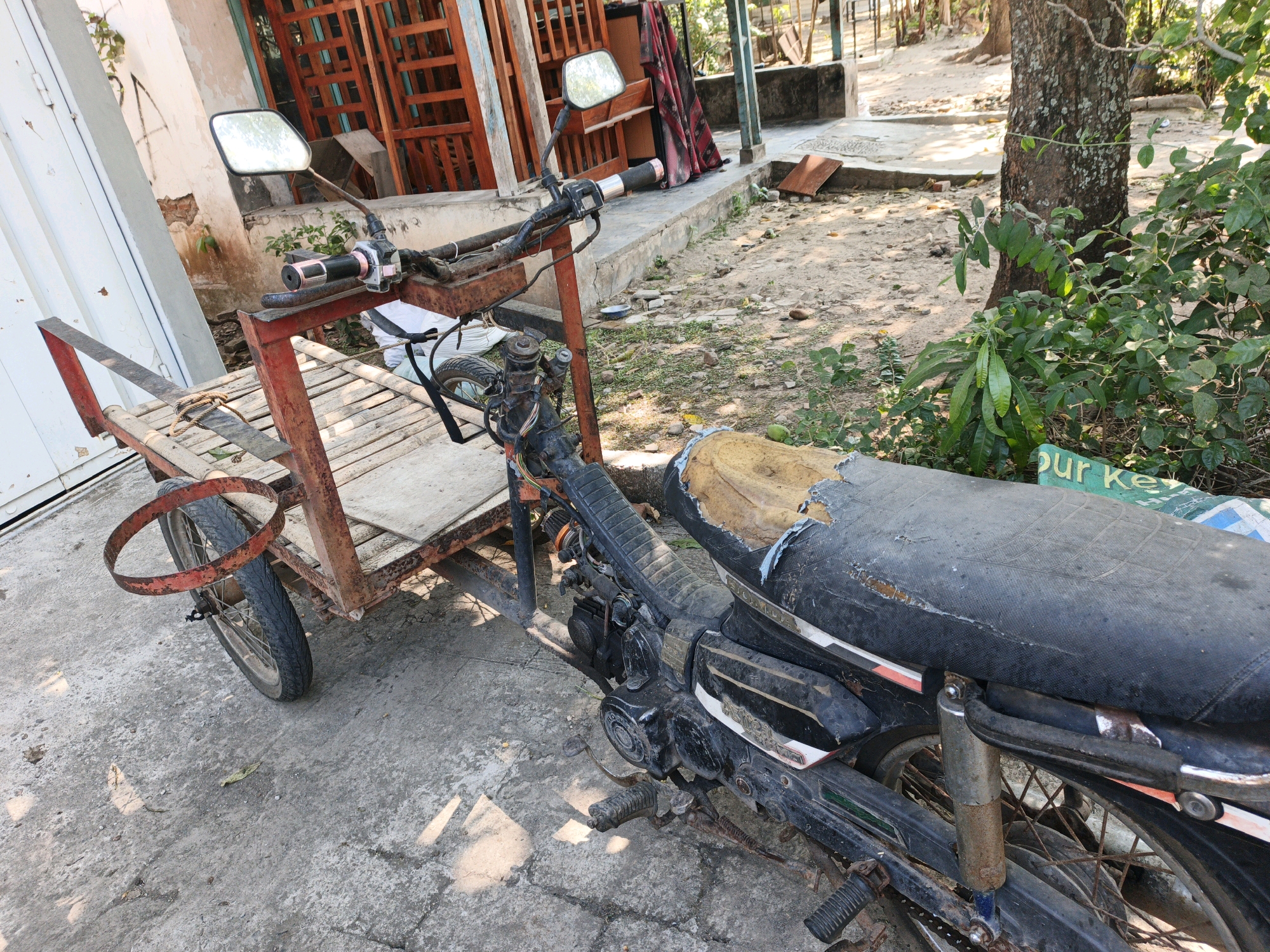 A three-wheeled motorized cart, constructed from a motorcycle and a trailer, is parked on a concrete surface outside a building in Modongan, Indonesia. The motorcycle's frame is painted black, and its seat is torn and weathered. The trailer, also showing signs of wear, is made of red metal and wood. The cart is surrounded by dirt, scattered leaves, and vegetation. In the background, there are wooden structures and trees, suggesting an outdoor, possibly rural or semi-rural, setting. The lighting indicates it is daytime.