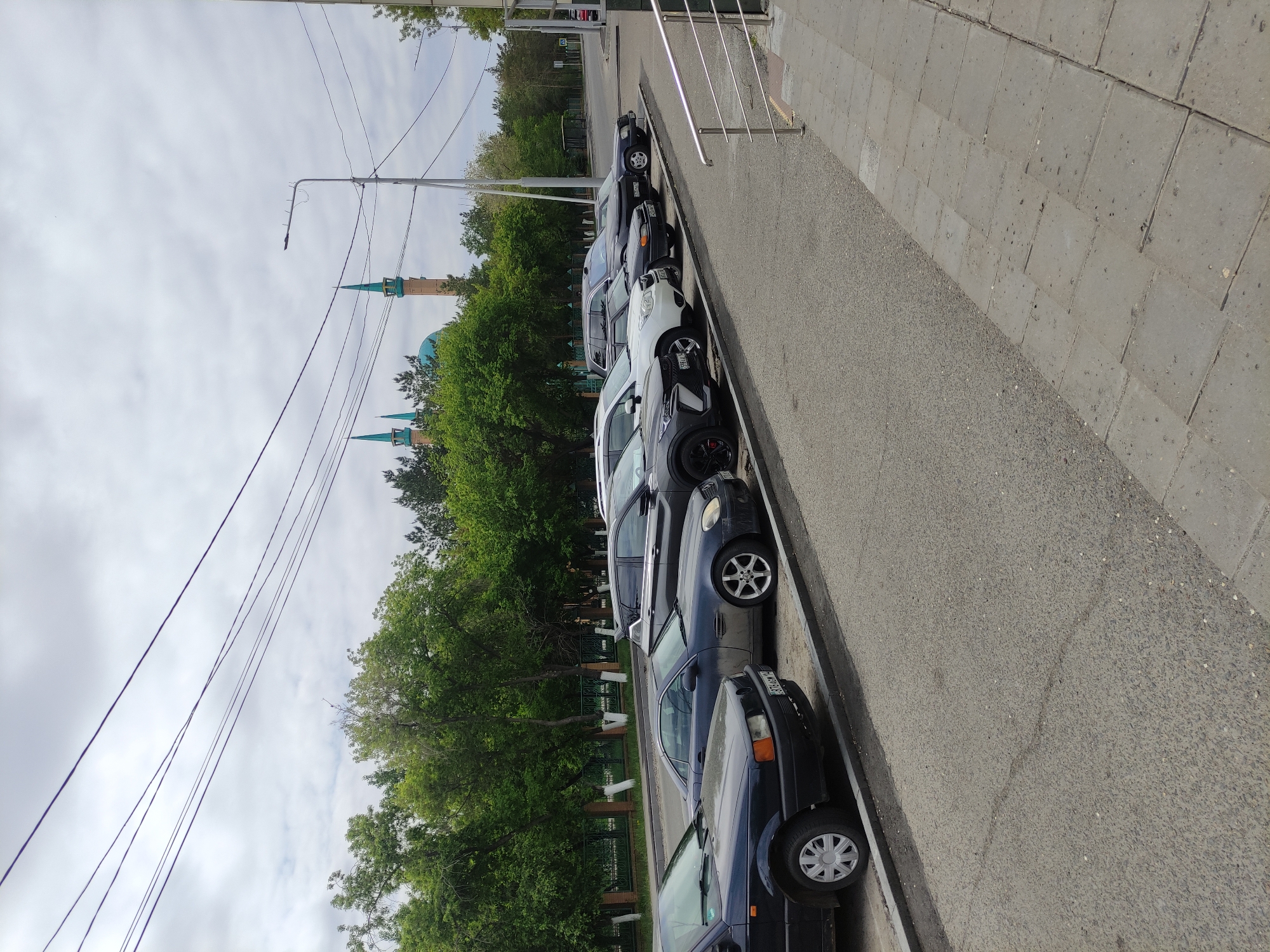 The image depicts a row of parked cars along a street in Pavlodar, Kazakhstan. The street is paved, with a sidewalk on the right side of the image, delineated by a metal railing. The vehicles are parked adjacent to a line of trees. Behind the trees, a building with turquoise domes and spires is visible. Overhead are electrical wires and a street light. The sky is cloudy. No people are visible in the frame.