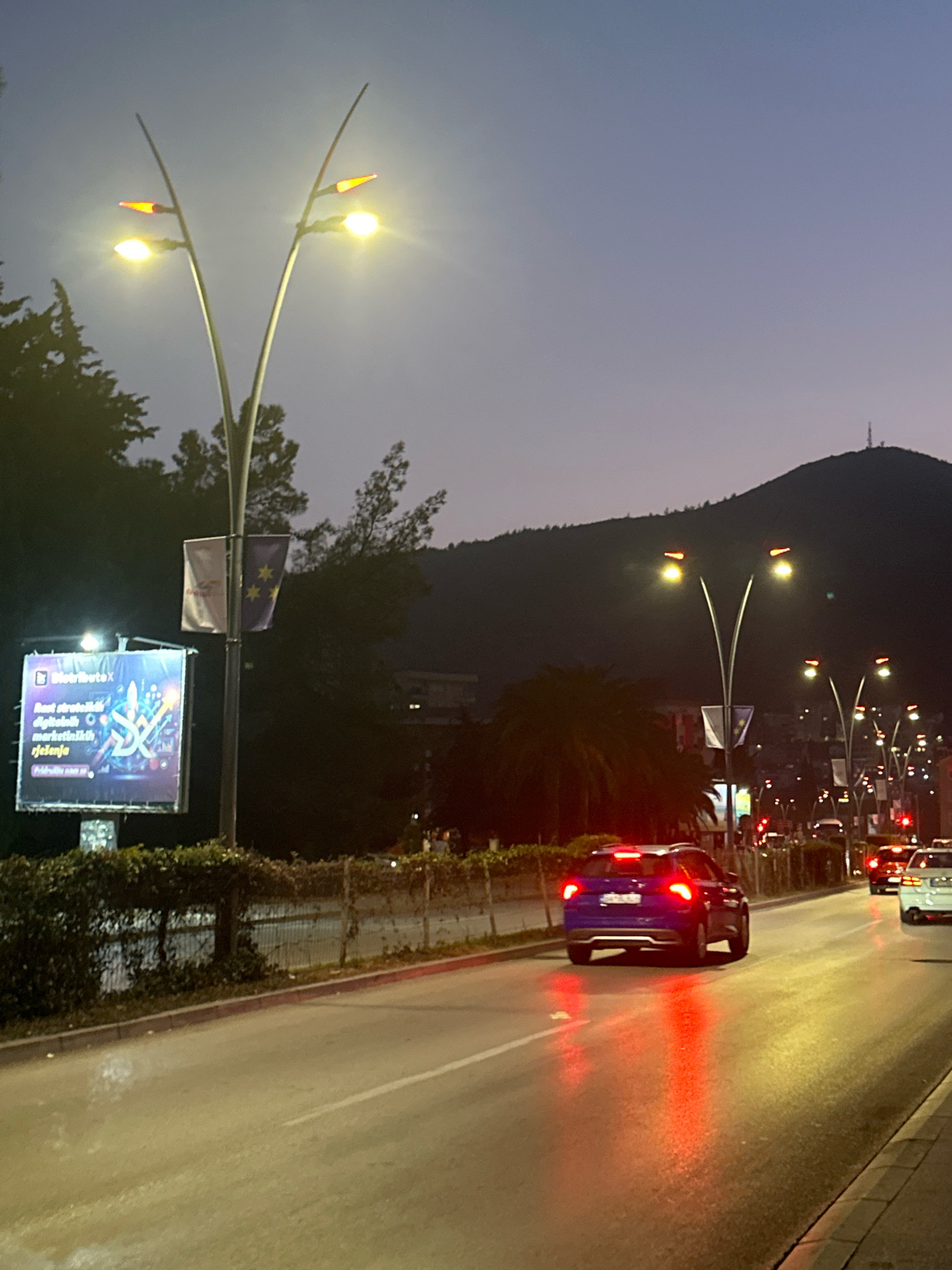 This is a nighttime, eye-level, outdoor shot taken on a road in the city of Budva, Montenegro. The foreground shows a asphalt road with two lanes, illuminated by streetlights and car headlights. A blue SUV is in the right lane, its brake lights on. To the left of the road, there is a sidewalk with a low hedge. Behind the hedge is a large billboard displaying an advertisement. To the left of the billboard, a tall, modern streetlight with two curved arms holds two lamps, one emitting a warm yellow light and the other a reddish-orange light. Next to the streetlight are two flags, one white and one purple with yellow stars. In the middle ground, more streetlights line the road, and palm trees are visible. In the background, a dark, sloped mountain dominates the skyline, with a small structure on its peak that appears to be a communication tower. Several buildings with illuminated windows are visible on the lower slopes of the mountain. The sky is a gradient of dark purple to a lighter, dusky blue. The overall scene depicts a street in Budva during twilight or early evening.