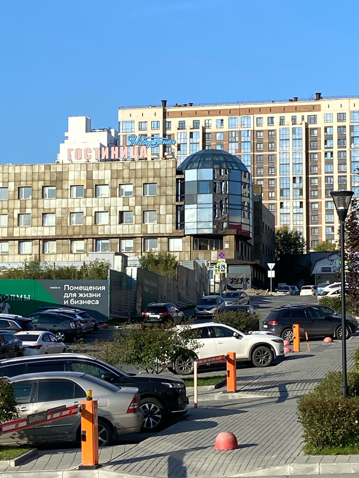 The image depicts a parking lot with several cars in various states of being parked. In the background, there are multiple buildings, some of which appear to be apartment complexes or office buildings. The tallest building on the right is a tan-colored multi-story structure with numerous windows. To the left of it is a building with a distinctive glass-domed section. A pink neon sign on one of the buildings reads "ГОСТИНИЦА" (Gostinitsa), which translates to "Hotel" in English. A green construction fence with white text indicating "Помещения для жизни и бизнеса" (Premises for life and business) is visible. The scene appears to be in an urban environment, likely a city, based on the architecture and presence of parked vehicles. The sky is clear and blue.