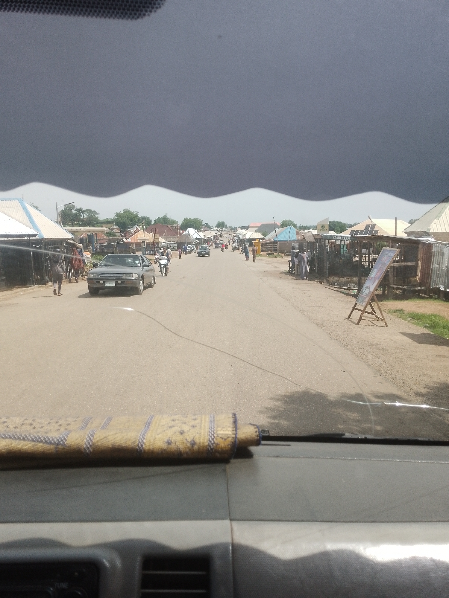 The image captures a street scene in Beli, Nigeria, likely during the daytime, given the illumination. The sky is dominated by a large, dark, and ominous cloud formation that spans the upper portion of the frame, with a distinctly wavy lower edge. This suggests impending or ongoing stormy weather.
The street is paved and appears to be a main thoroughfare in a town or village. On either side of the road are numerous small shops and market stalls, many with corrugated metal roofs. Some stalls have awnings or overhangs. The architecture suggests a local, possibly informal, commercial area.
Several people are visible, walking along the road and near the stalls. A few individuals are dressed in traditional attire, including flowing robes. There are also motorcycles and a car on the road, indicating regular traffic. One car, a sedan, is prominent in the foreground on the left side of the road. The presence of a few scattered trees indicates some vegetation in the area.
In the foreground, obscuring the bottom of the image, is the dashboard of a vehicle, with a textured material draped over a part of it. This suggests the photo was taken from inside a car, looking out through the windshield.
On the right side of the road, a wooden easel displays a sign with some text and imagery, possibly an advertisement or information board. The overall atmosphere is that of a busy, though potentially unsettled, day in a Nigerian community.
Visible text includes "LIMITED" on a structure in the background and partially visible text on the sign on the easel. The license plate on the car is also visible but unreadable.
