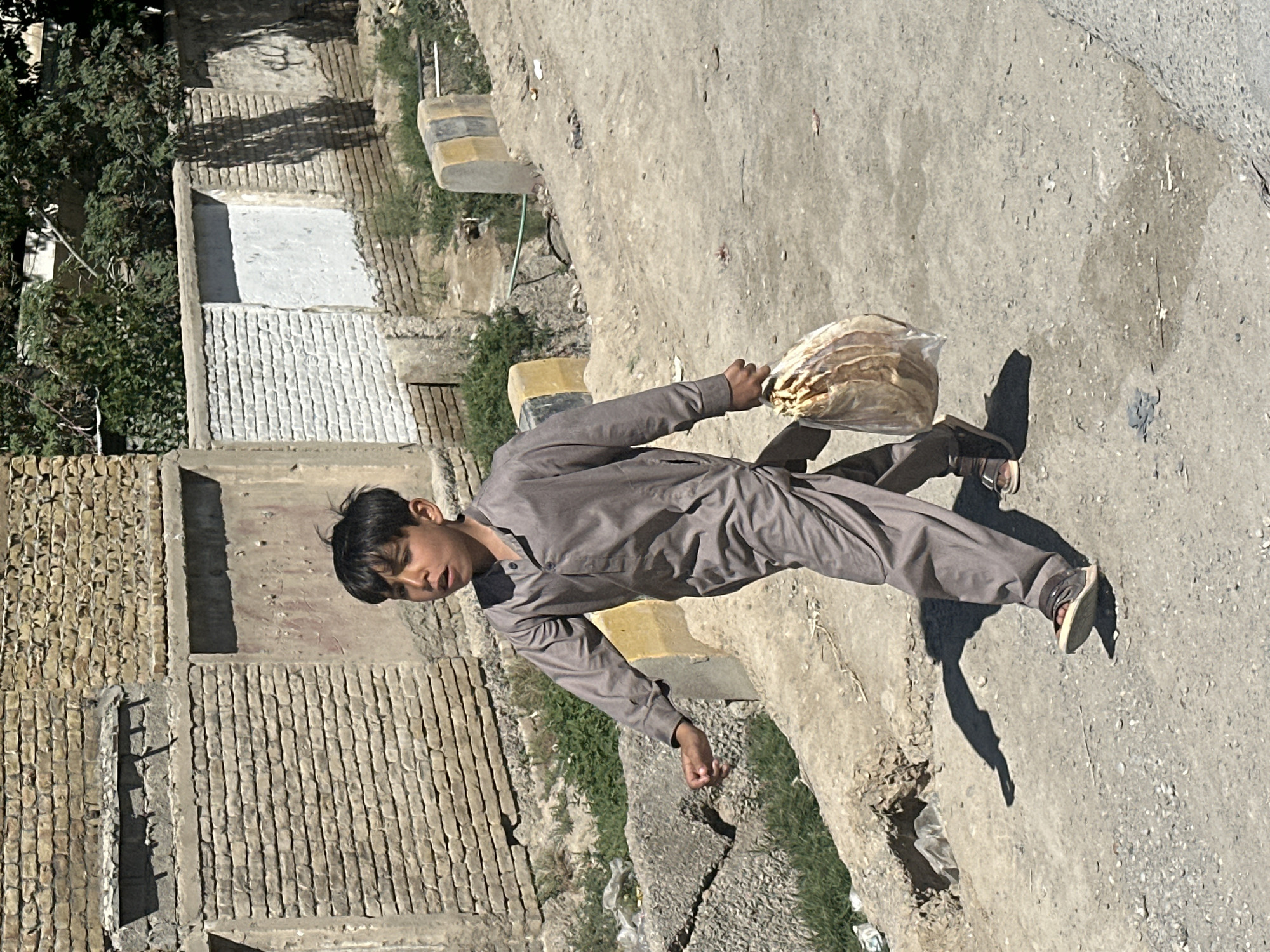 A young boy is walking along a paved road in what appears to be the Quetta City Tehsil, Pakistan. He is wearing a grayish-brown, long-sleeved jumpsuit and shoes. The boy is carrying a clear plastic bag filled with flatbread. The background features buildings constructed from tan brick, with some vegetation growing along the sides of the road. The sunlight suggests it's daytime. The scene has a dusty and arid feel.
