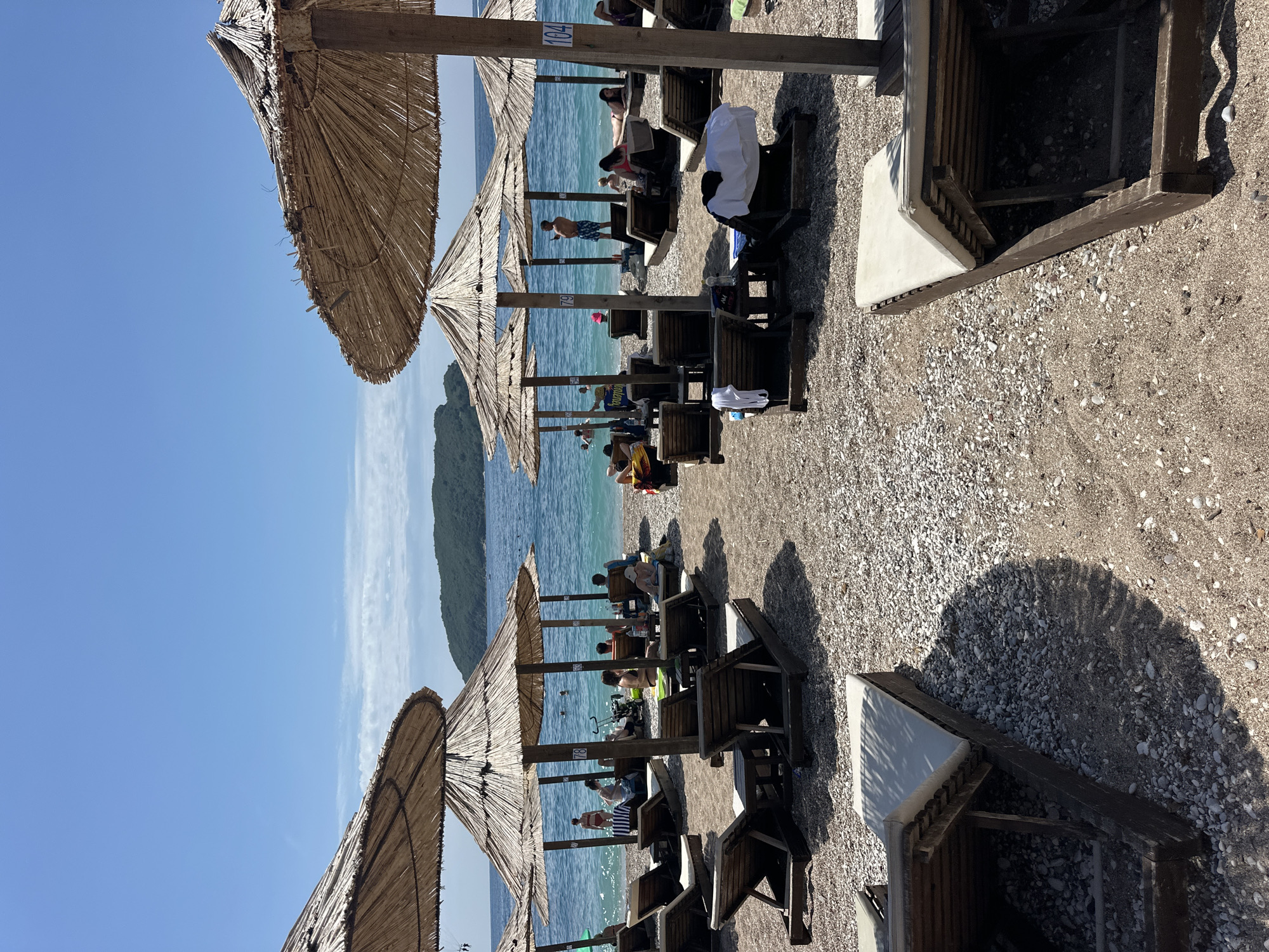 The image depicts a vibrant and sunny beach scene in Budva, Montenegro, bustling with people enjoying a clear day. The foreground shows a pebbly or gravelly beach lined with rows of traditional straw beach umbrellas, casting prominent shadows onto the ground. Beneath these umbrellas are wooden sun loungers, some fitted with white cushions, occupied by individuals relaxing.
Numerous people are visible across the beach and in the water. Some are reclining on the loungers, while others are swimming or standing in the clear, blue-green sea. One person can be seen on a blue inflatable that has the word "Gulliver" printed on it. In the background, a serene blue sky with a few wispy clouds meets the horizon, beyond which a lush, green, hilly landmass or island rises from the water.
The bright sunlight and strong shadows suggest it is likely midday or early afternoon. Visible text includes the numbers "104" and "89" marked on the wooden poles of two different beach umbrellas, likely indicating their station or rental number. The overall atmosphere is one of leisure and vacation at a popular coastal destination.