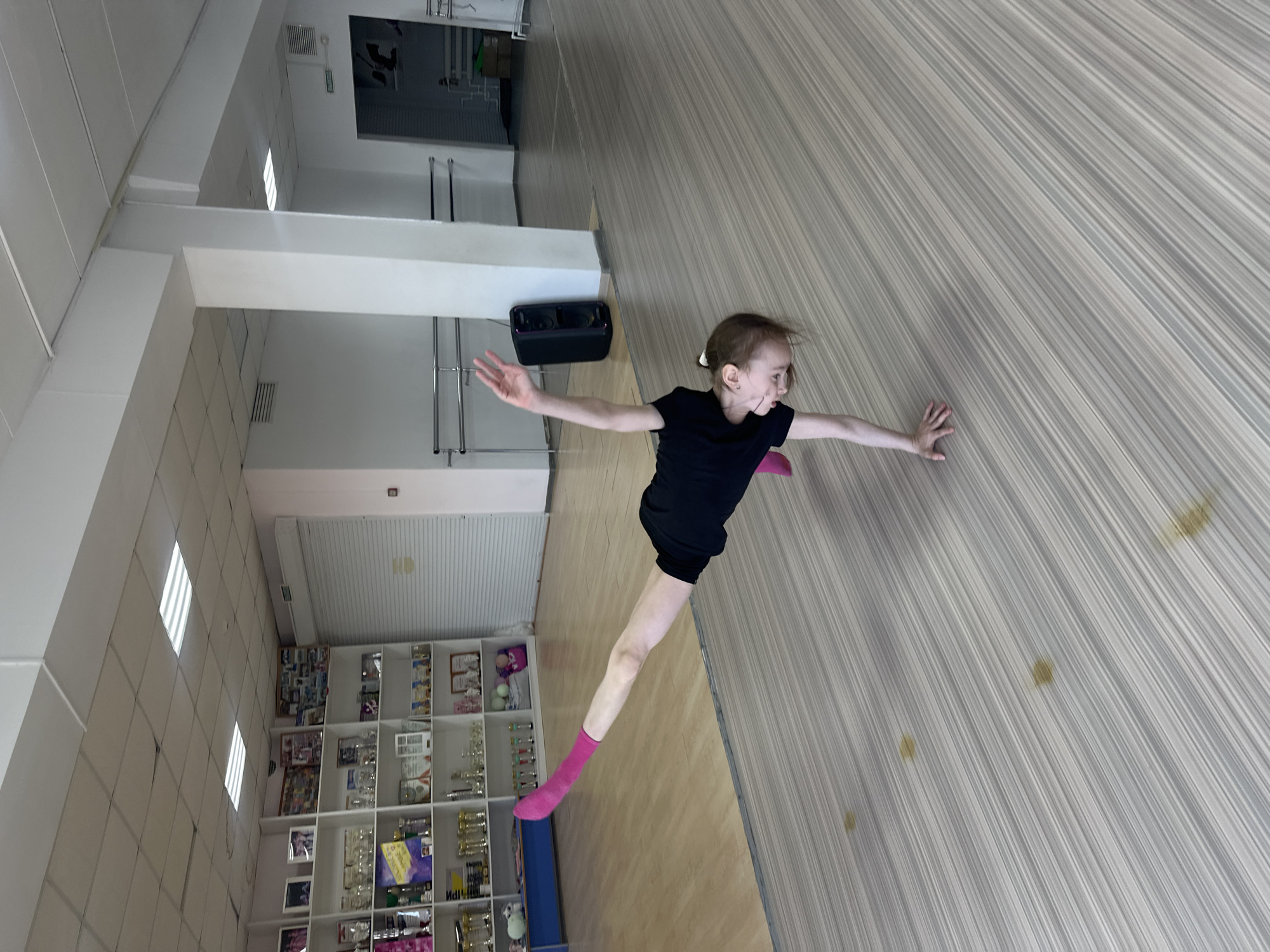 A young female child wearing a black leotard and pink socks is striking a dance pose in a studio in Votkinsk, Russia. The studio has a light-colored striped floor, white walls, and a display cabinet filled with various objects, including awards and trophies. There's a black speaker on a shelf. The child has her arms extended to the sides.