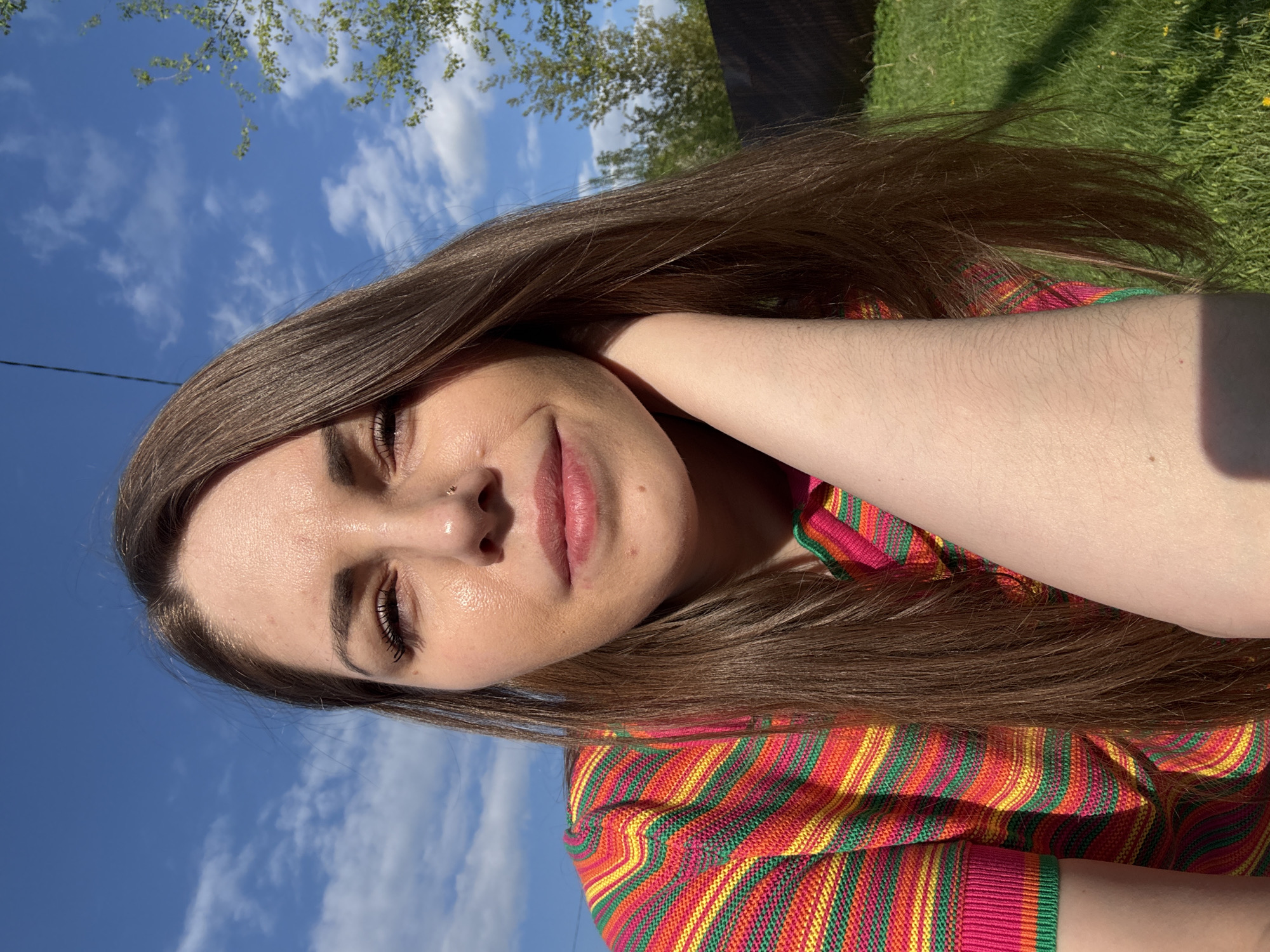 The image shows a selfie of a woman with long brown hair, taken outdoors in daylight. She is wearing a colorful striped shirt with pink, red, green, and yellow hues. Her head is slightly tilted, and her right arm is raised with her hand touching her neck. The background features a blue sky with scattered white clouds and green foliage of trees, suggesting a natural setting. In the upper portion of the background there is also green grass. Given the prompt's request to include a location in the city of Раздолье, Russia, the image could theoretically be set in a park or outdoor recreational area within that city.