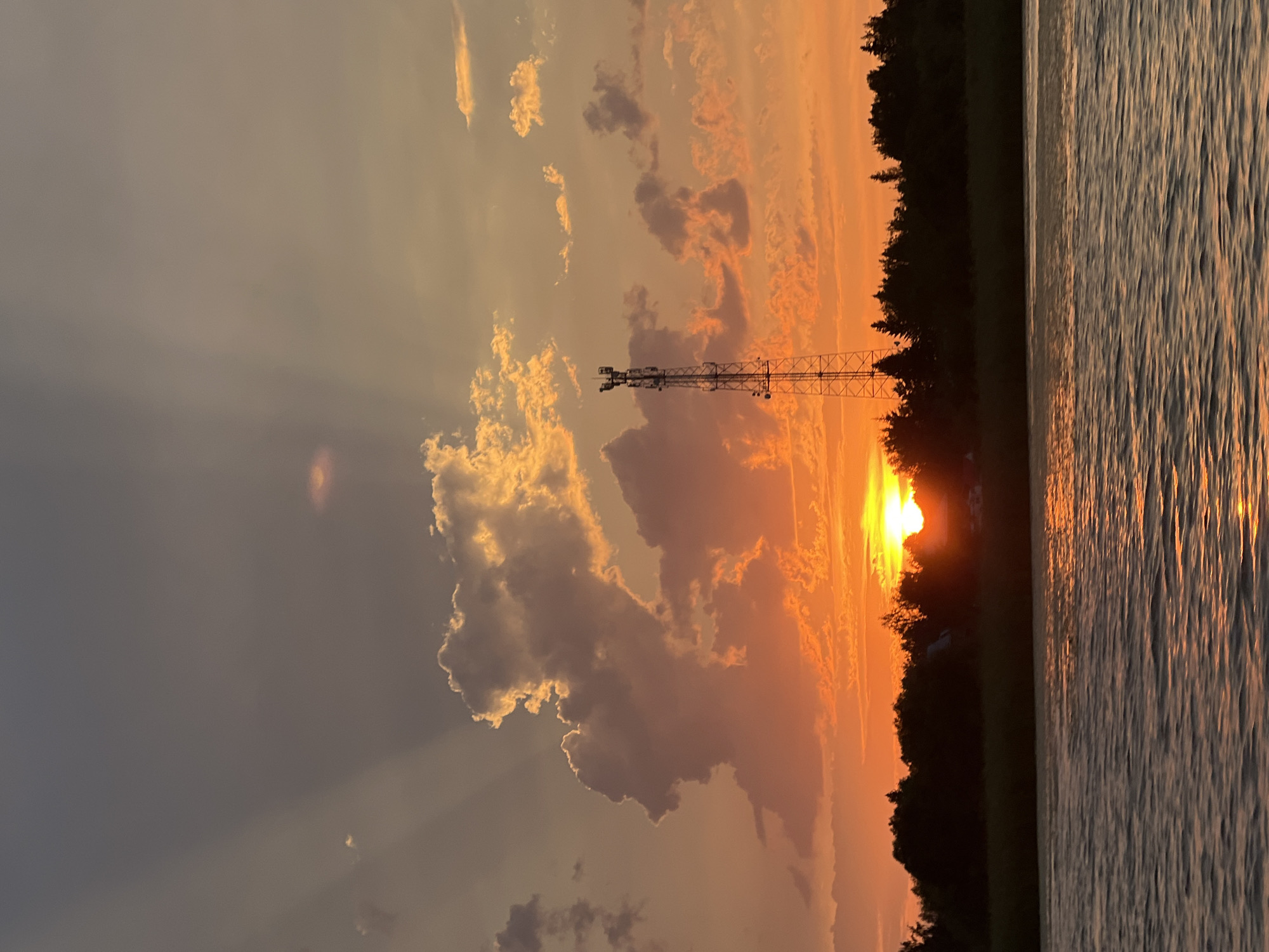 The photograph captures a sunset over a body of water, likely a river or lake, in Novopichugovo, Russia. The sky is filled with a dramatic display of color, ranging from deep orange and yellow near the horizon to cooler grays and purples higher up. Sun rays pierce through scattered clouds, creating a luminous effect. A tall, metal telecommunications tower stands prominently in the middle ground, silhouetted against the vibrant sky. A dense line of dark trees forms the shoreline, with a few glimpses of structures, possibly houses, nestled within the foliage. The water in the foreground reflects the warm hues of the sunset, with gentle ripples creating a textured surface. There are no people or animals visible in the scene.