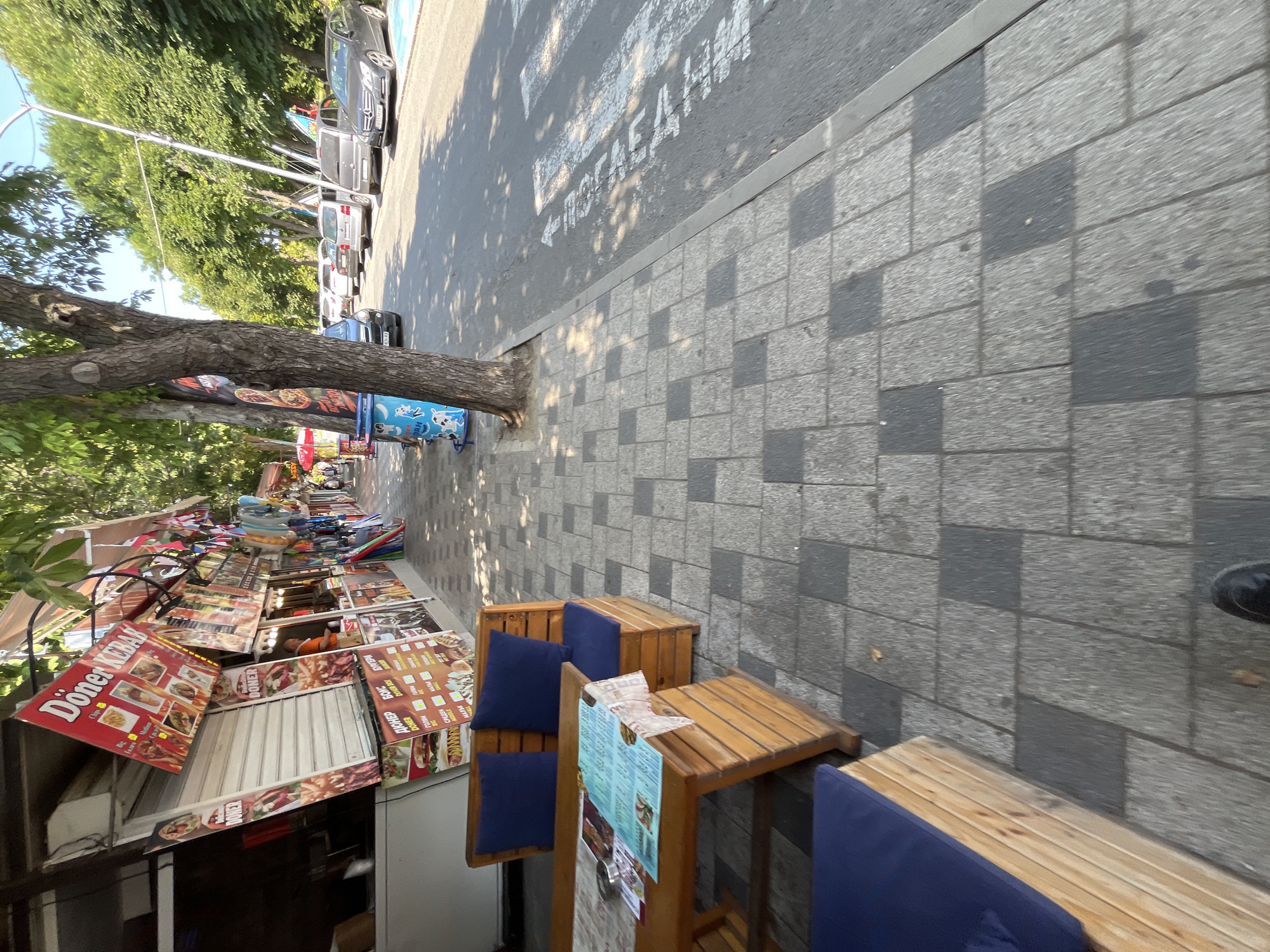 The image captures a street scene in Obzor, Bulgaria, likely a commercial area with several food vendors and a sidewalk. In the foreground, a wooden table and bench with blue cushions are set up, suggesting an outdoor seating area for a restaurant or cafe. Menus are spread across the table. The sidewalk is paved with interlocking grey and dark grey rectangular tiles. Beyond the seating area, a row of food stalls lines the street, displaying signs in Cyrillic and English advertising "Döner Kebab," "Burger," and other food items. Colorful flags and decorations are visible above the stalls. A large tree with dense green foliage provides shade. In the background, cars are parked along the street, and a pedestrian crossing is marked on the asphalt. The environment appears to be a sunny day, with sunlight casting dappled shadows on the ground.