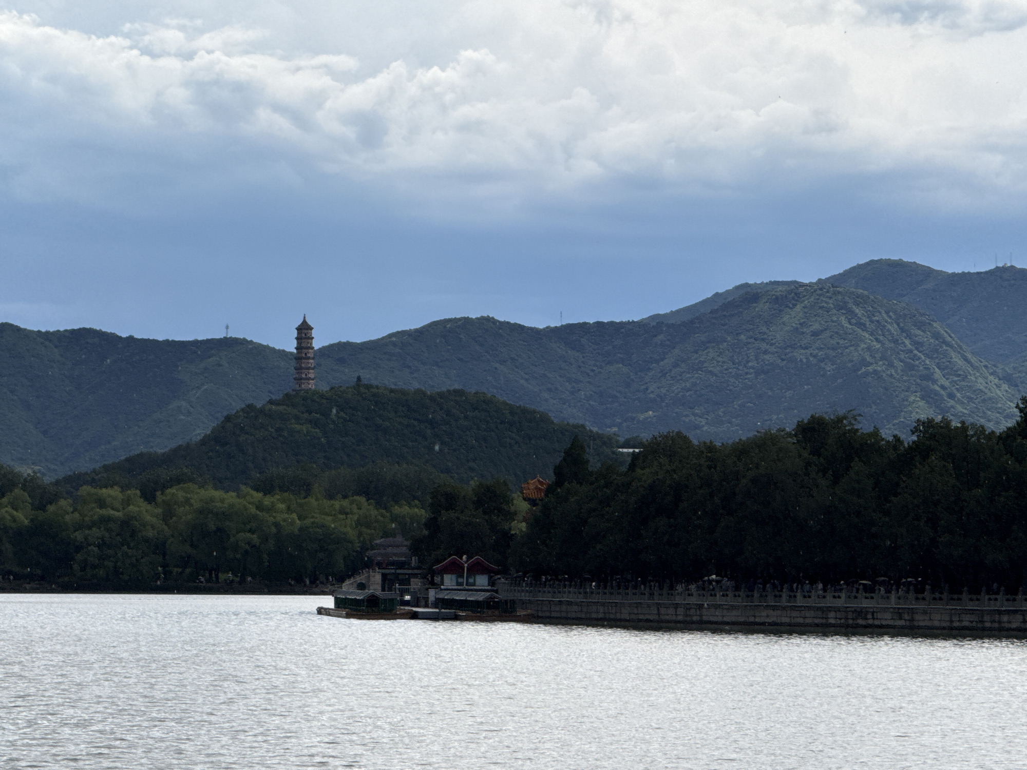 This forensic description details a wide landscape photograph taken in Haidian District, China, depicting a large body of water in the foreground with forested hills and architectural elements in the midground and background under an overcast sky.
The foreground is occupied by a calm, greyish-white body of water, showing fine ripples across its surface. In the midground, along the right shoreline, a long, ornamental stone balustrade or railing runs horizontally. Behind this balustrade, a dense line of dark green trees obscures much of the land, though indistinct figures of people are visible walking along a path or promenade. Further left along the shoreline, several small structures with red-tiled roofs and lighter-colored walls are situated at the water's edge, accompanied by a few small boats moored nearby.
The background consists of multiple layers of rolling hills, uniformly covered in dense green foliage. These hills recede into the distance, appearing progressively lighter due to atmospheric perspective. On the most prominent hill, positioned towards the left-center, a tall, multi-tiered pagoda with a distinct architectural style is visible. Another slender, antenna-like structure is faintly visible on a more distant hill to the right of the pagoda. The sky above is entirely overcast, dominated by a mix of light grey and blue-grey clouds.