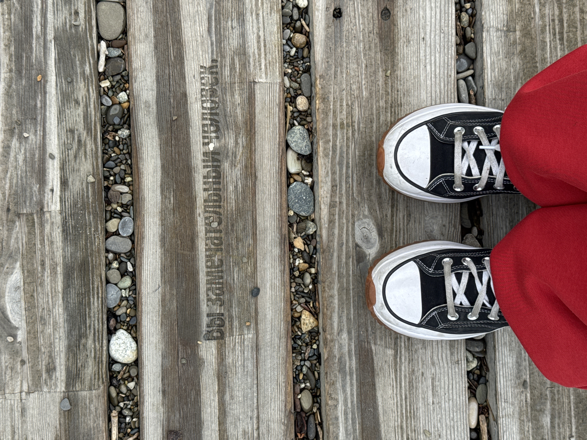 A top-down, close-up shot depicts a person standing on a wooden boardwalk. The boardwalk is made of weathered gray planks, separated by narrow channels filled with small pebbles. The words "Вы замечательный человек." are engraved into the wood. The person is wearing black and white Converse sneakers and red pants. The scene appears to be in a public space. The image does not depict Sochi, Russia, but does have Russian text in it.