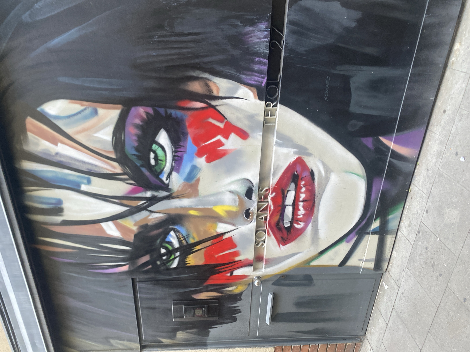 The image displays a vibrant mural on a dark metal surface, possibly a garage door or a building facade, located on Terol 27 in Barcelona, Spain. The mural is a striking portrait of a woman's face, rendered in bold, expressive strokes and a dynamic color palette. The woman's eyes are a piercing green, emphasized by colorful makeup, including shades of purple, blue, and red. Her lips are painted a vivid, glossy red, with her teeth slightly visible. The artwork features loose, brush-like strokes in black, white, and various colors that create a sense of movement and energy, particularly in her hair, which appears to be dark and choppy. There are also touches of yellow and brown on her face, adding to the abstract and emotive quality of the portrait. Below the face, a metallic plaque or sign reads "SOLANES" in silver lettering, and further to the right, the street number "TEROL 27" is also visible in similar lettering, confirming the location. Another smaller signature, "SOLANES," is subtly placed on the dark background to the right of the main portrait. The mural is situated outdoors, with a gray tiled sidewalk in the foreground. A small, dark metal intercom or electrical box is attached to the left side of the painted surface, and a gray handle, possibly for a door, is positioned below it. The overall impression is of a striking piece of street art that adds a bold artistic statement to its urban environment.