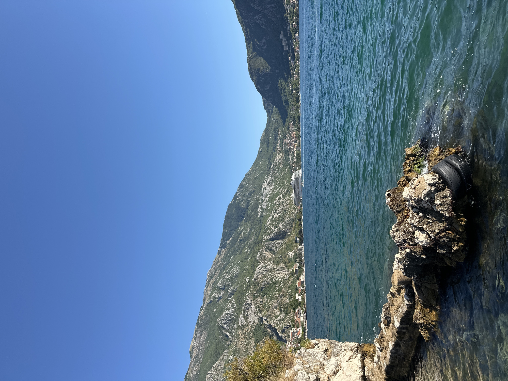 The image shows a body of water, likely the Bay of Kotor, with a town nestled along the shoreline in the background. Steep, verdant mountains rise behind the town. A large cruise ship is visible in the distance, as are some smaller boats. In the foreground, a rocky shoreline juts into the water. The water is a deep blue-green, and the sky is clear and bright blue. The scene appears to be a scenic coastal view.