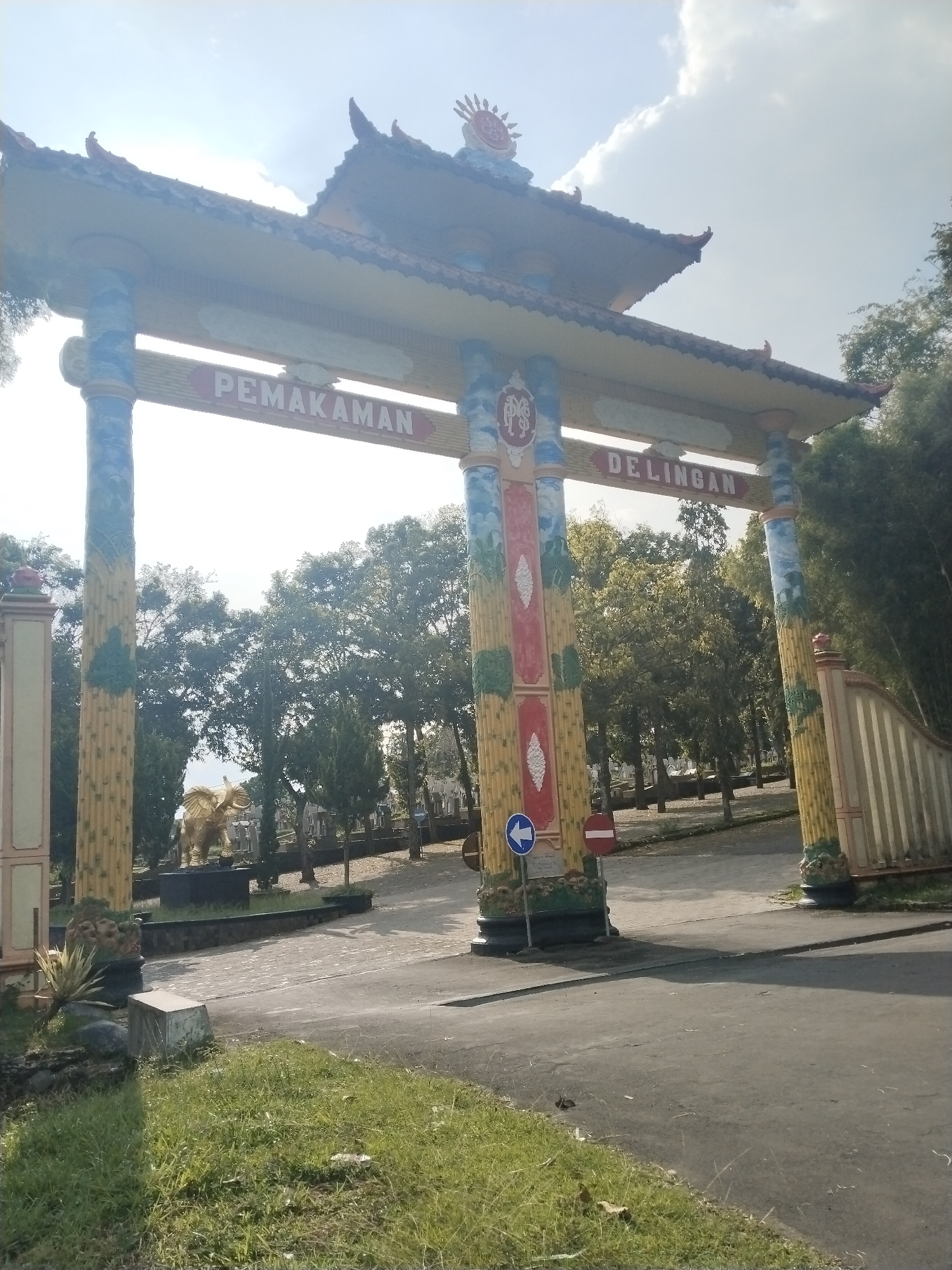 The image captures a grand, ornate entrance gate under a bright, sunny sky with scattered clouds. The gate features a multi-tiered, pagoda-style roof and is supported by four large, elaborately decorated pillars. The pillars are painted in vibrant colors, primarily yellow with green and blue accents, resembling bamboo stalks and foliage.
Across the main archway of the gate, large red letters spell out "PEMAKAMAN DELINGAN." Below this text, on the central pillars, a red emblem with white intertwined letters, possibly "P" and "D" or "M" and "S", is visible. To the left of the left central pillar, a blue circular traffic sign with a white left-pointing arrow is posted. Another red circular sign with a white horizontal bar (indicating no entry or stop) is visible to the right of the same pillar.
Beyond the gate, a paved road leads into a spacious area filled with numerous tall, leafy trees, suggesting a park or a large open ground. Amongst the trees and set back from the road, a prominent golden statue of an elephant can be seen. On the far right, a white fence or wall borders the area. The foreground features patchy green grass and a paved section of road leading up to the gate. No people are visible in the scene. The bright lighting and clear shadows indicate it is daytime, likely mid-morning or early afternoon. The overall setting is the entrance to the Delingan Cemetery in Ngernak Lor, Indonesia.