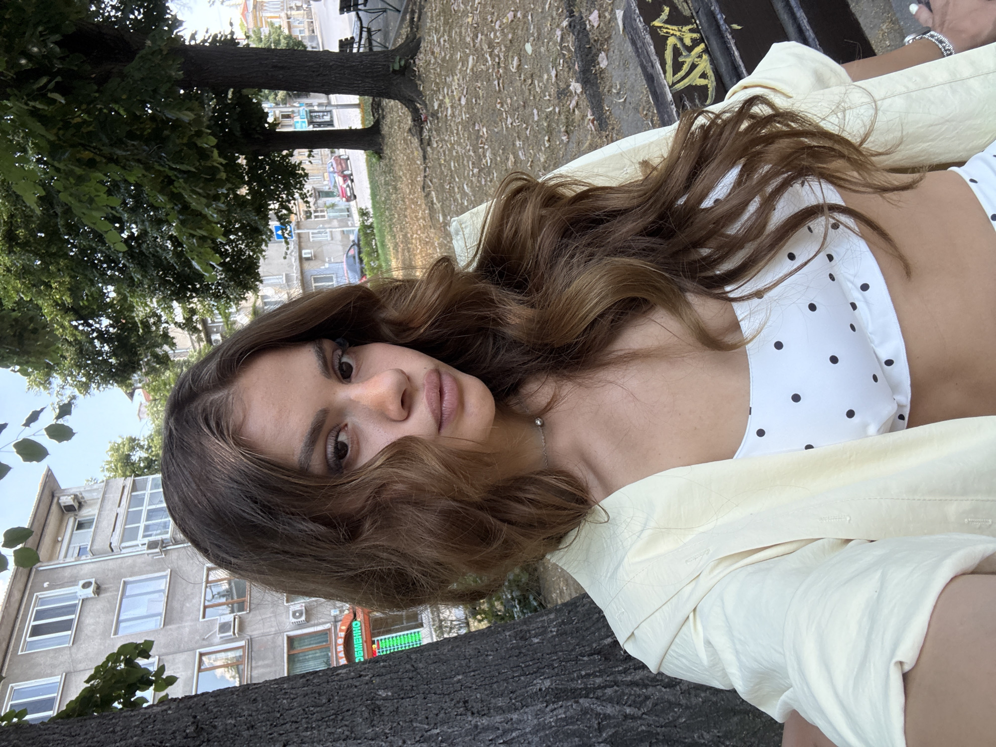 The media file shows a portrait of a young woman lying on her back, looking up at the camera. She has long, wavy brown hair and is wearing a white polka dot top and a light yellow open jacket. Her gaze is direct and her lips are slightly parted.
The setting appears to be outdoors, possibly a park or a public seating area. In the background, there are trees with green foliage and fallen leaves scattered on the ground. Beyond the trees, a residential building with multiple windows and air conditioning units is visible. The sky is bright, suggesting daytime. The overall mood seems relaxed and casual.
There is some text visible on a sign in the background, which reads "ЗЕМЕДЕЛИЕ" (Zemedelie), meaning "Agriculture" in Bulgarian. The location context is identified as Plovdiv, Bulgaria. The image has a slightly tilted perspective, as if taken from a low angle.