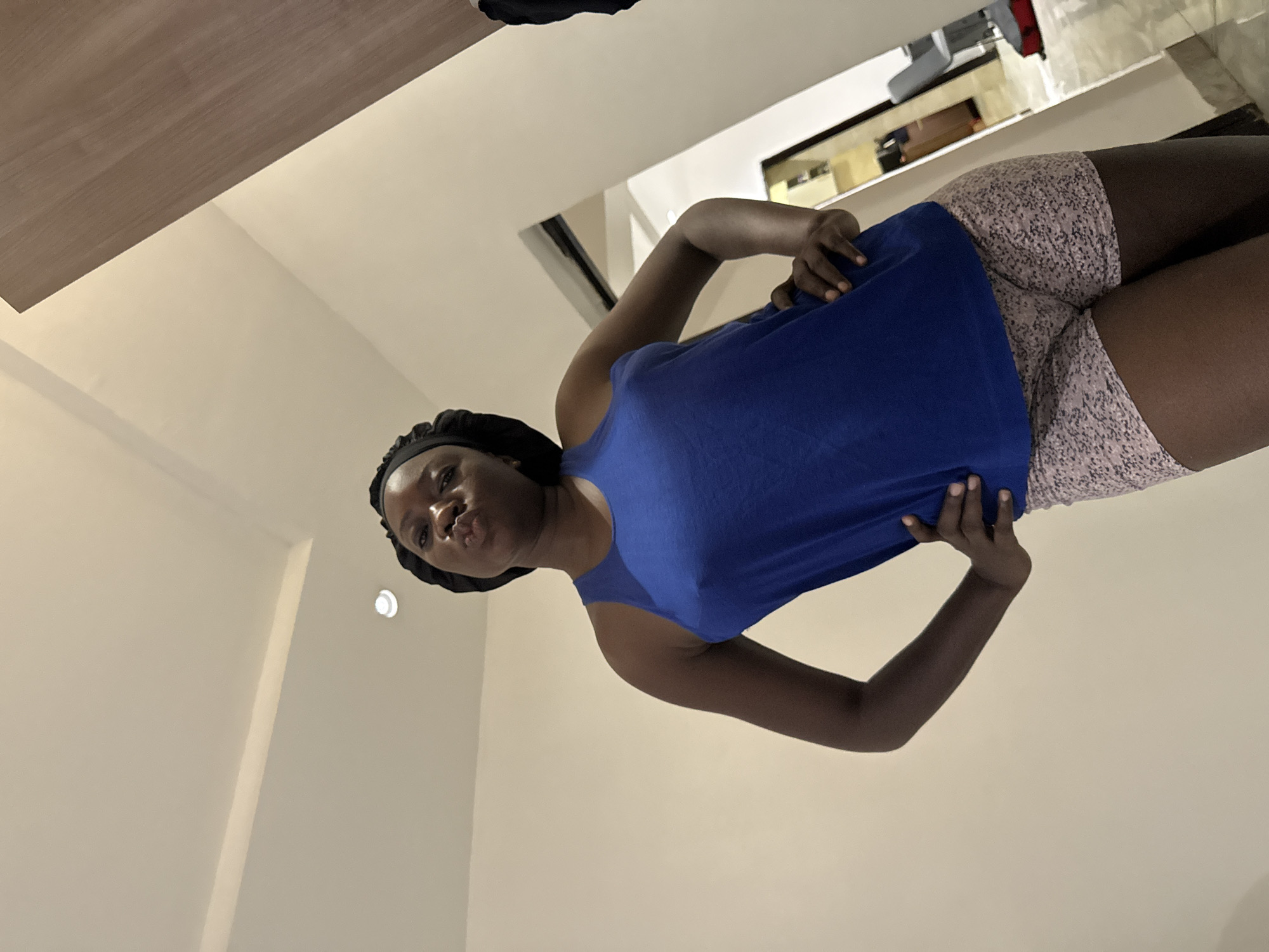 An indoor, slightly low-angle, full-shot captures a woman posing playfully. She stands with her body angled slightly left, hands on her hips/waist, and looks directly at the camera with a pouting expression. She is wearing a sleeveless royal blue top and light-colored, possibly pink or white, patterned shorts. A black head covering or scarf is visible on her head.
The background consists of light-colored, possibly off-white or beige, walls. Above her head, a wooden shelf or ceiling section is visible. To her right, a reflective surface, likely a mirror or glossy furniture, shows a blurred reflection of another part of the interior. A small, circular light fixture is visible on the wall behind and to her left. The setting suggests a modern indoor space within a residence or building in Iddo Tudunwada, Nigeria, illuminated by artificial lighting or diffused daylight. No text is visible in the image.