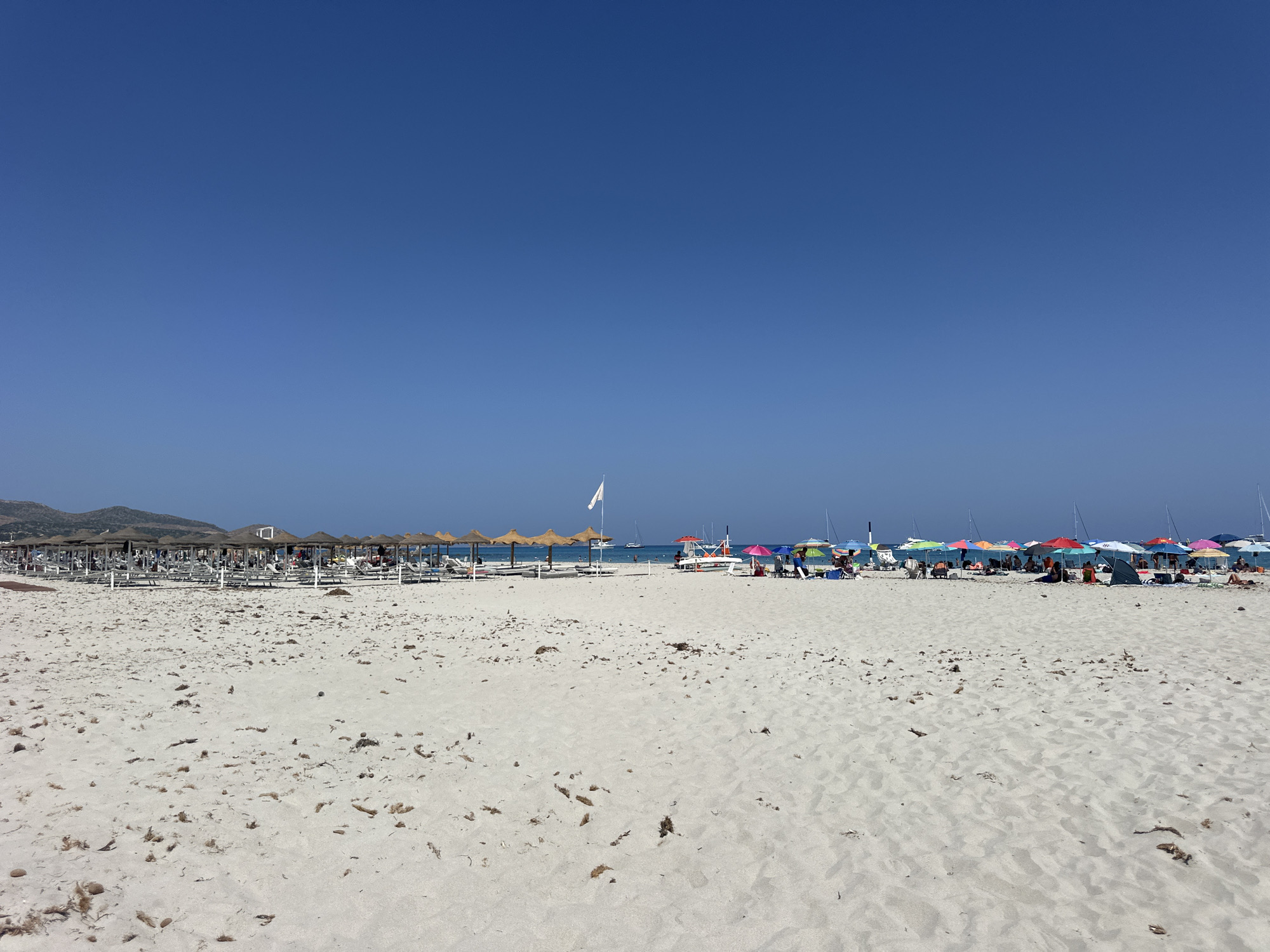 This is a wide-angle, eye-level photograph of a sandy beach in Crabonaxa, Villasimius, Italy. The foreground is dominated by white sand with scattered dried seaweed and debris. In the middle ground, a row of thatched-roof umbrellas and lounge chairs stretches across the beach, with a few people visible relaxing. Beyond this setup, a calm sea is visible, dotted with sailboats. The background features a clear, bright blue sky. The overall scene depicts a typical summer day at a popular Italian beach resort.