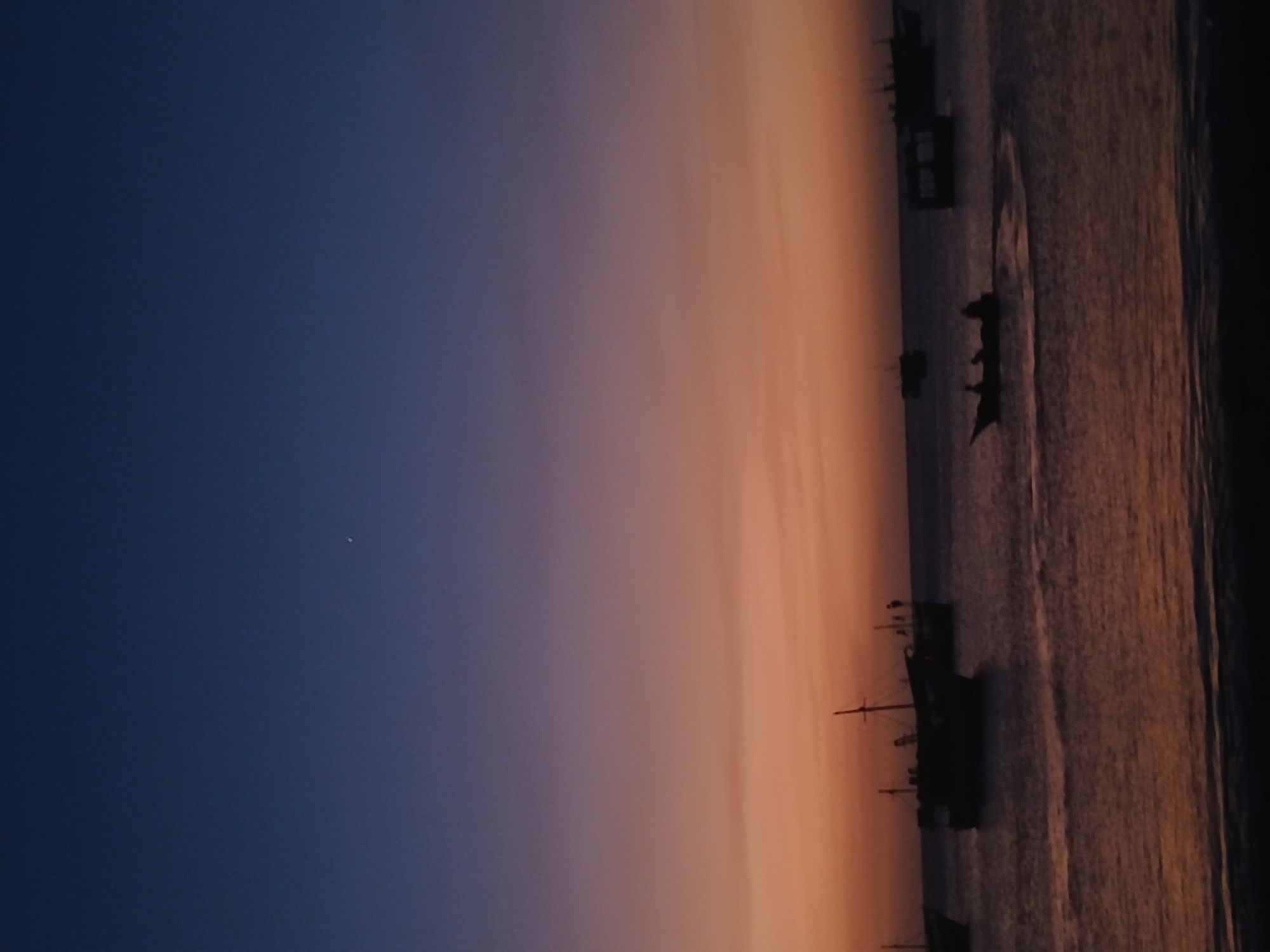 The image depicts a waterfront scene at dusk in Juan Griego, Venezuela. The sky transitions from dark blue in the upper left to orange near the horizon. Several silhouetted boats are visible on the water near the right edge of the image. A small point of light is present in the dark blue sky.