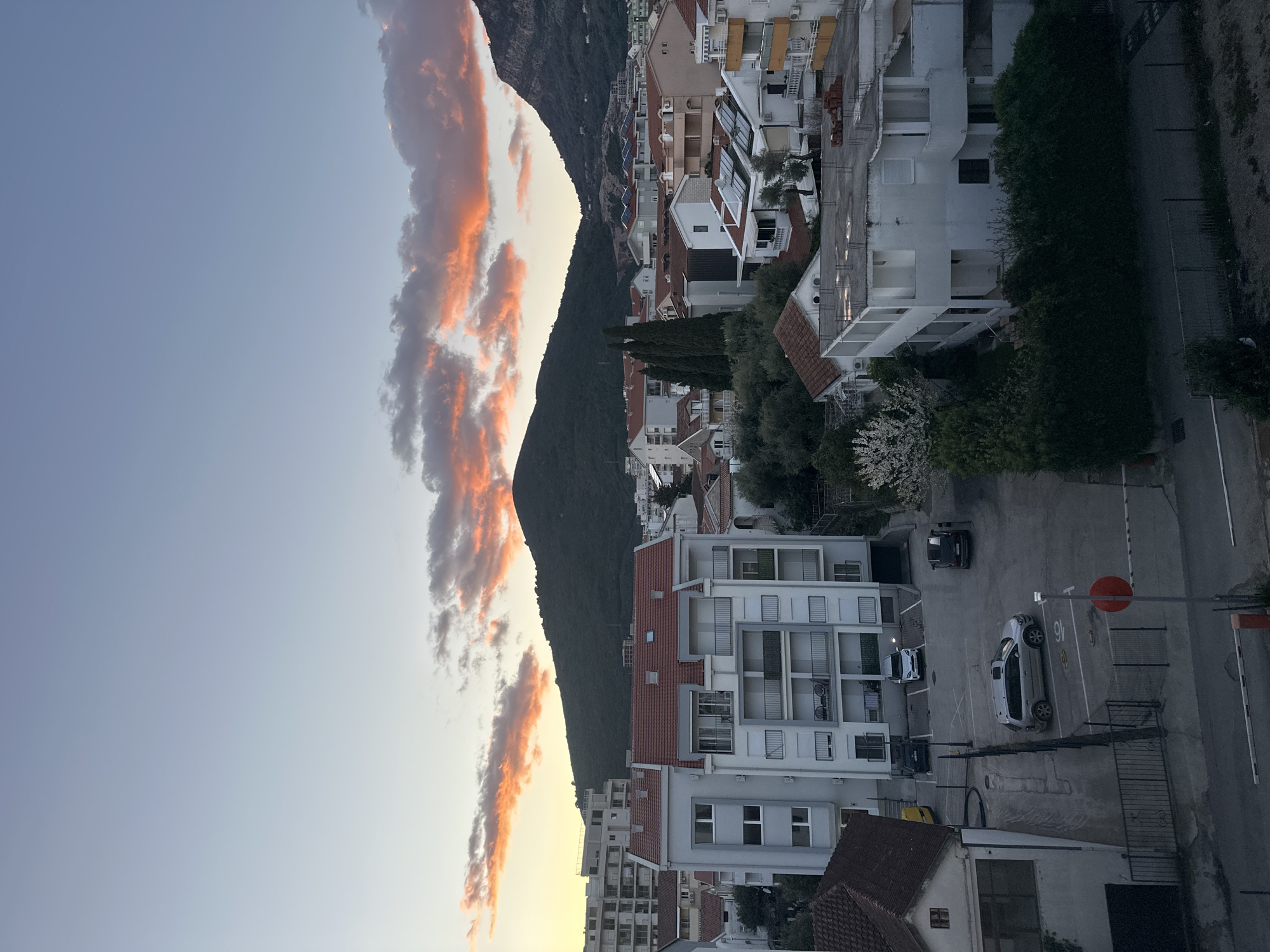 The image depicts a view of Budva, Montenegro at sunset. The sky is visible with orange and pink clouds reflecting the setting sun. A dark, forested mountain rises vertically in the center. To the right of the mountain is a dense collection of white and light-colored buildings with red-tiled roofs, indicative of a residential area. The buildings appear to be multi-story apartment complexes. In the lower right portion of the image is a concrete parking area with a few parked cars. A stop sign is visible on a pole next to the parking area. The overall scene is captured from an elevated perspective.