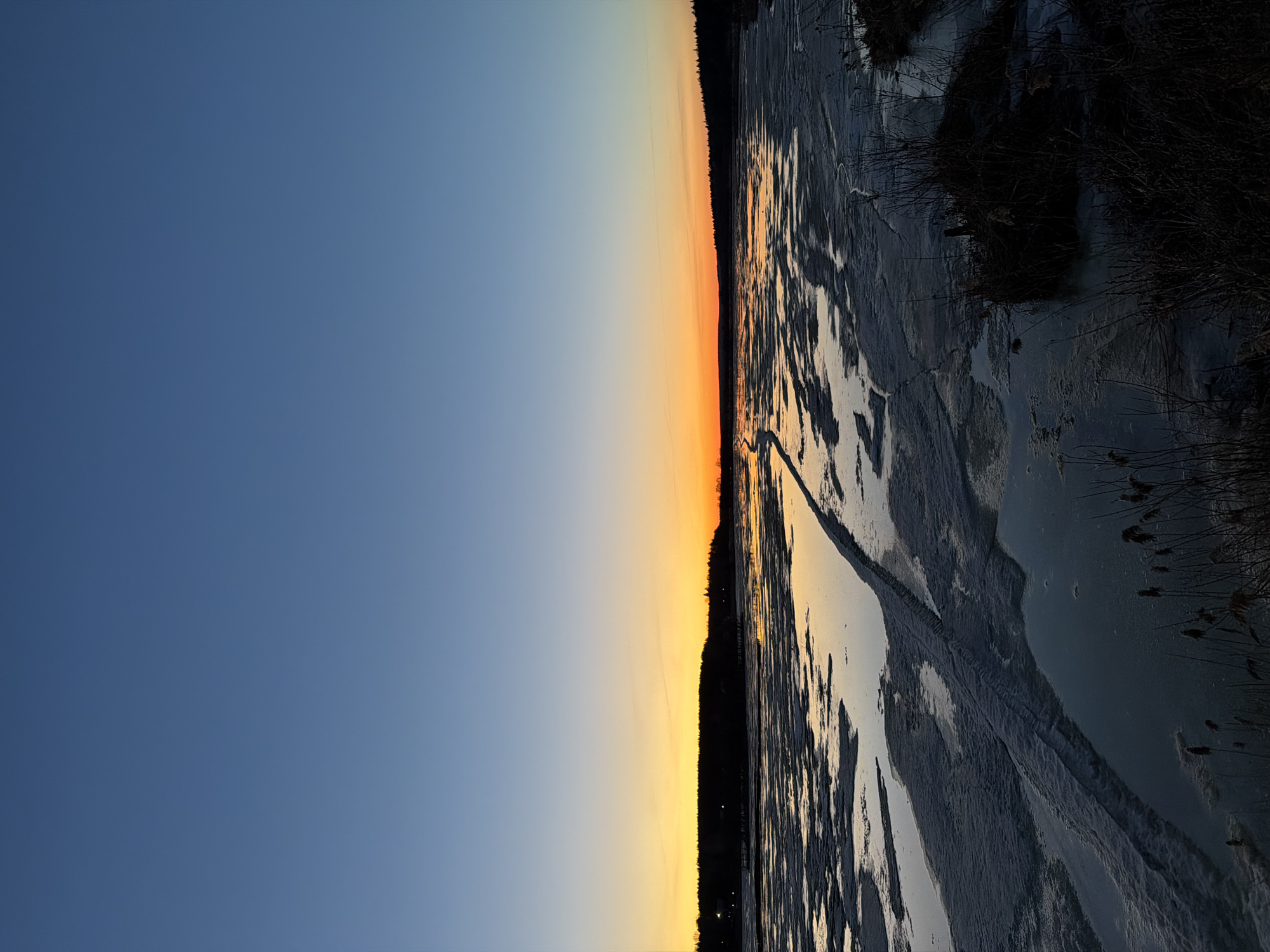 The image shows a winter scene during sunset in Izhevsk, Russia. The left side of the image depicts a clear, blue sky transitioning into a vibrant orange and yellow gradient at the horizon. The right side captures a frozen body of water, likely a pond or a section of the Izhevsk reservoir, covered with ice and snow. Dark silhouettes of trees line the horizon, and some vegetation can be seen along the shoreline on the right. There are traces of tire tracks visible on the ice, suggesting vehicle passage or activity on the frozen surface. The light from the setting sun reflects on the ice, creating a mirrored effect that blends the colors of the sky with the frozen landscape. The composition is landscape, with a strong horizontal emphasis, emphasizing the transition between the sky and the frozen ground.