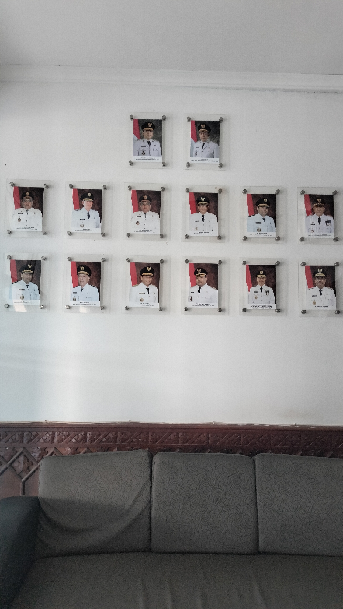 The image shows a wall with a series of framed portraits of men in formal attire, presumably officials. The portraits are arranged in three rows. The top row has three portraits, the middle row has five, and the bottom row has four. Each portrait is of a man wearing a white uniform with a black cap, and each has a small Indonesian flag on the left side of the frame. Below each portrait is a plaque with text, likely names and titles. The wall is white. Below the portraits, a dark brown wooden carved panel is visible, and beneath that, a dark gray sofa with textured cushions. The lighting appears to be ambient indoor lighting, casting soft shadows. The setting is likely a government office or public building in Lhokseumawe, Indonesia, given the context of the portraits and the Indonesian flag.